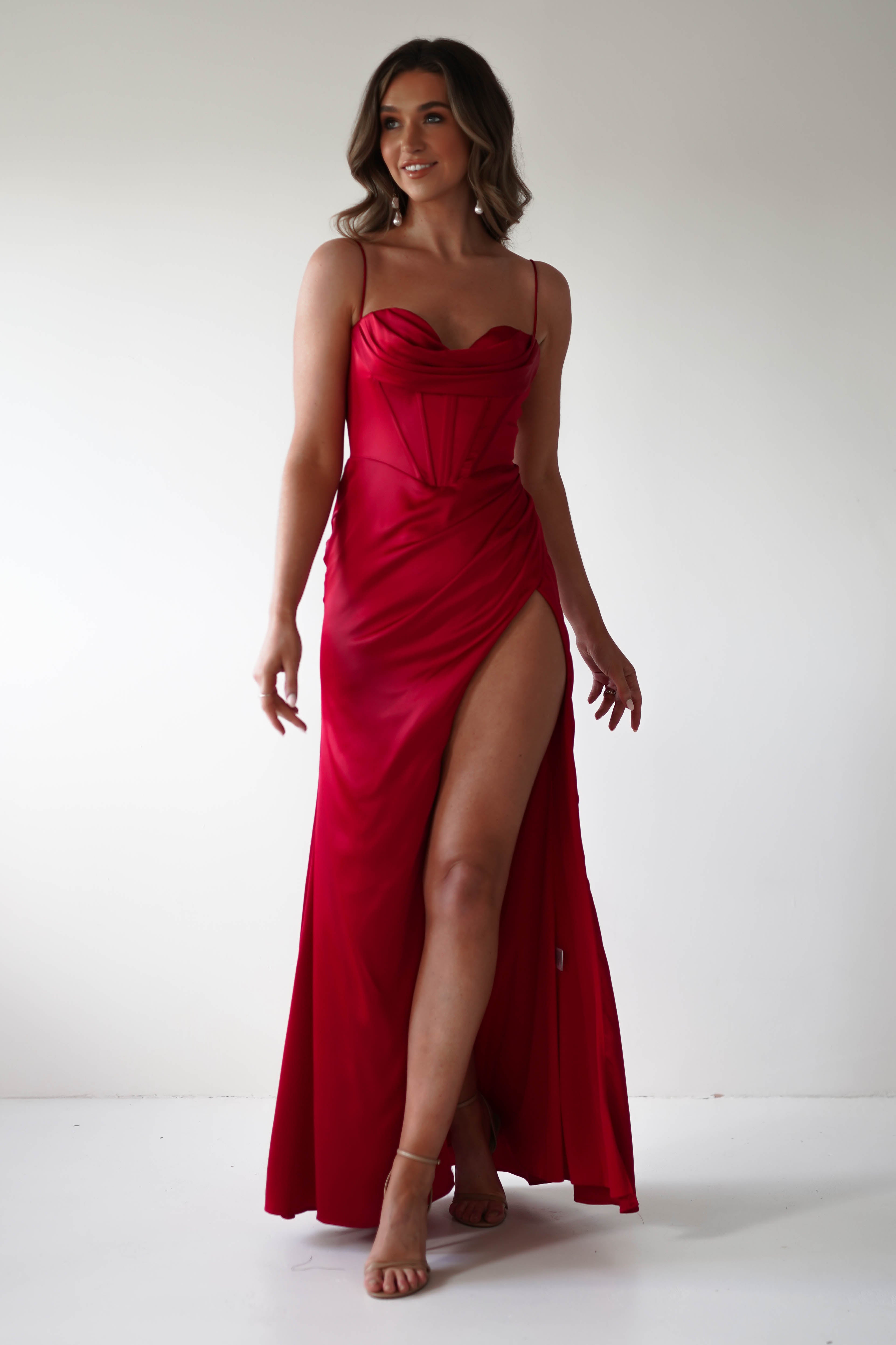 A woman in a stunning red corset gown with a high slit stands against a plain white background. She poses confidently with one leg forward, wearing strappy heels. Perfect for any special occasion, her soft waves complete the elegant look.