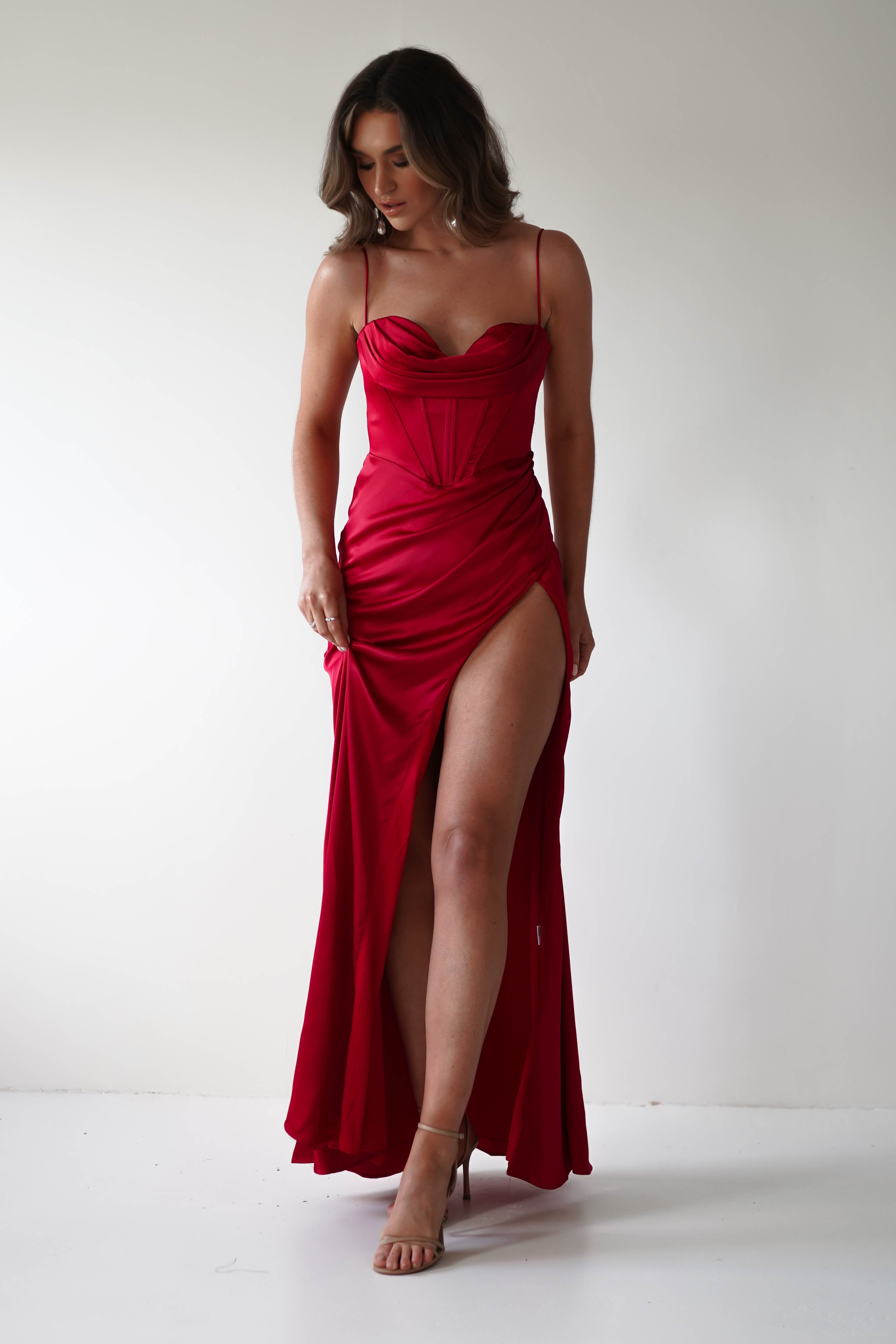 A woman poses against a plain background, wearing a sleeveless, form-fitting red corset gown with a thigh-high slit. She holds part of the dress with her left hand, revealing strappy high heels, perfect for any special occasion. Her hair is styled in loose waves.