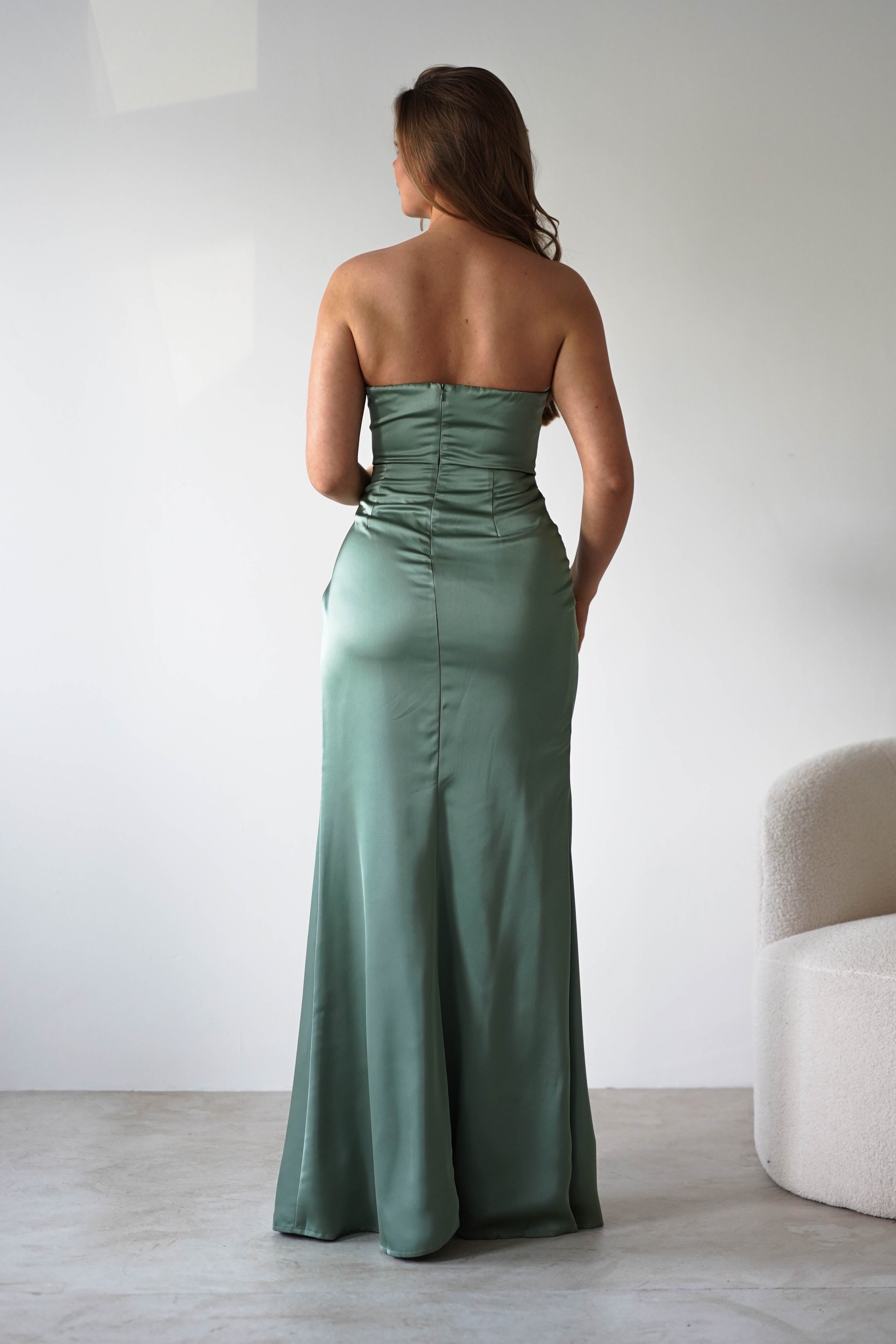 A woman in a strapless gown of sage green satin fabric stands with her back to the camera in a minimal, light-colored room with a modern, curved chair on the right—an elegant look perfect for any formal event.