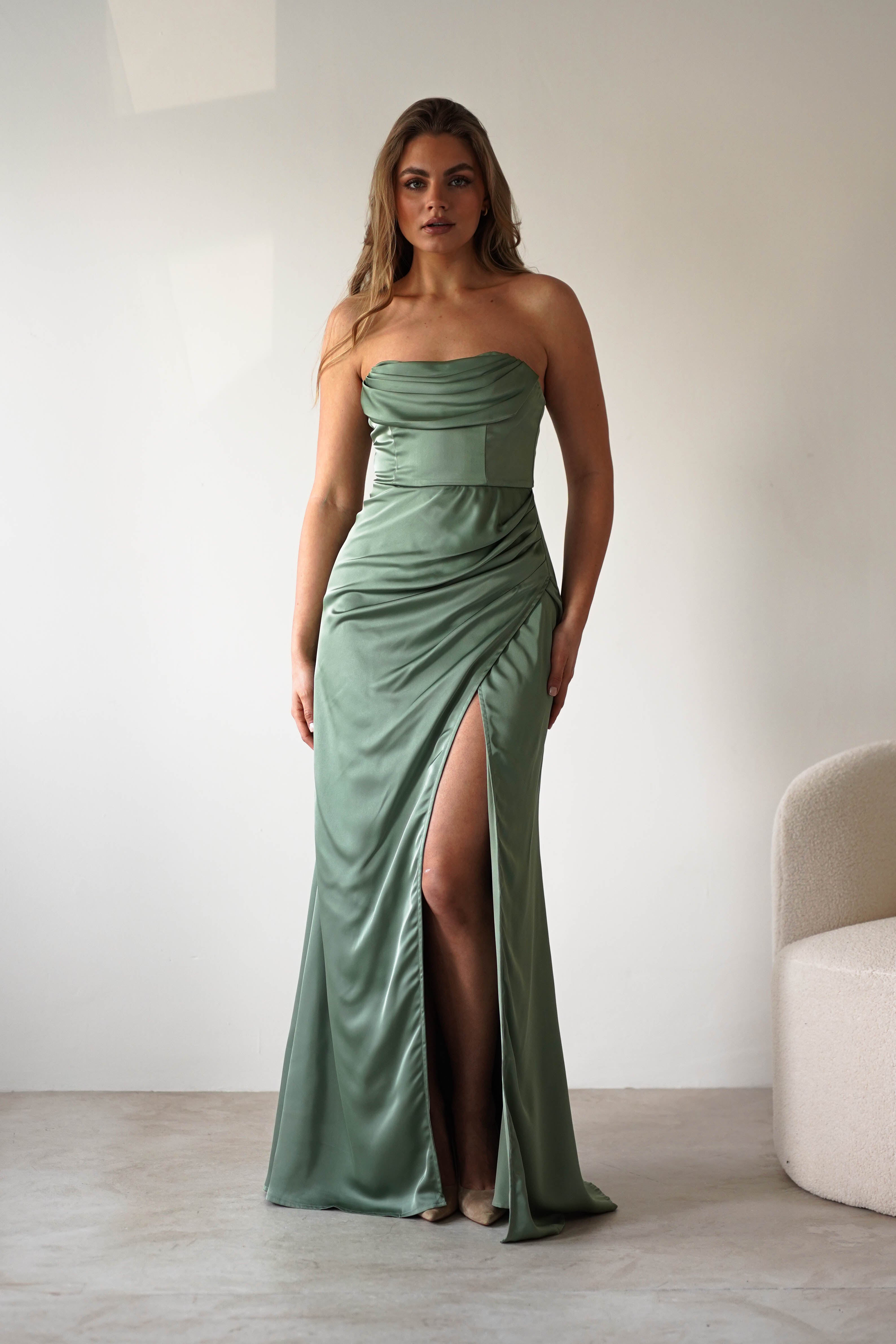 A woman in a strapless gown made of sage green satin fabric with a thigh-high slit stands indoors against a plain white wall next to a curved, light-colored chair.