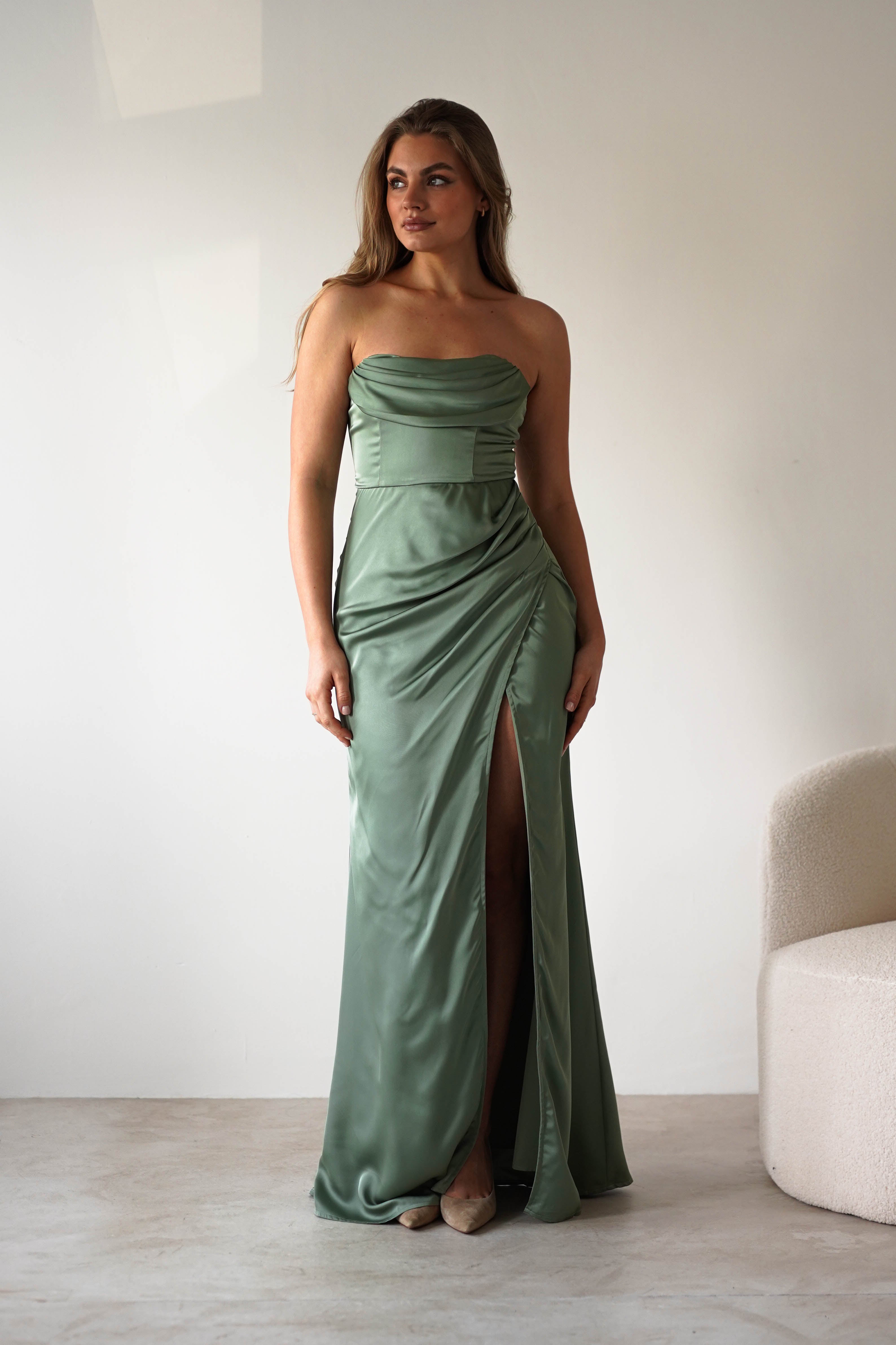 A woman in a light green, strapless satin gown with a thigh-high slit stands indoors on a light floor near a white, textured chair, looking slightly to her left—ready for a formal event.