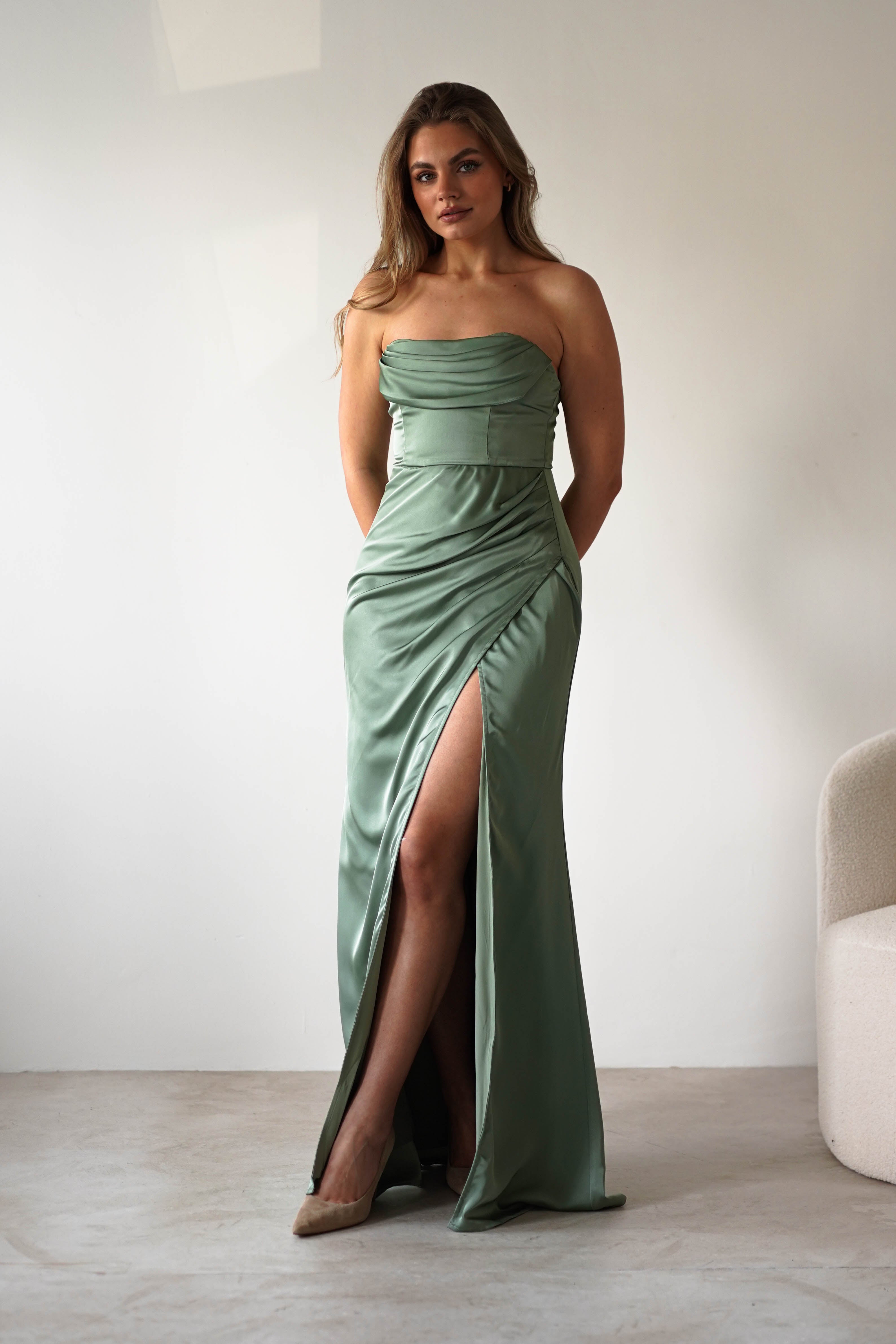A woman in a strapless satin gown with a thigh-high slit stands indoors on a light floor, her hands behind her back. She wears nude heels and looks confidently at the camera, ready for a formal event.