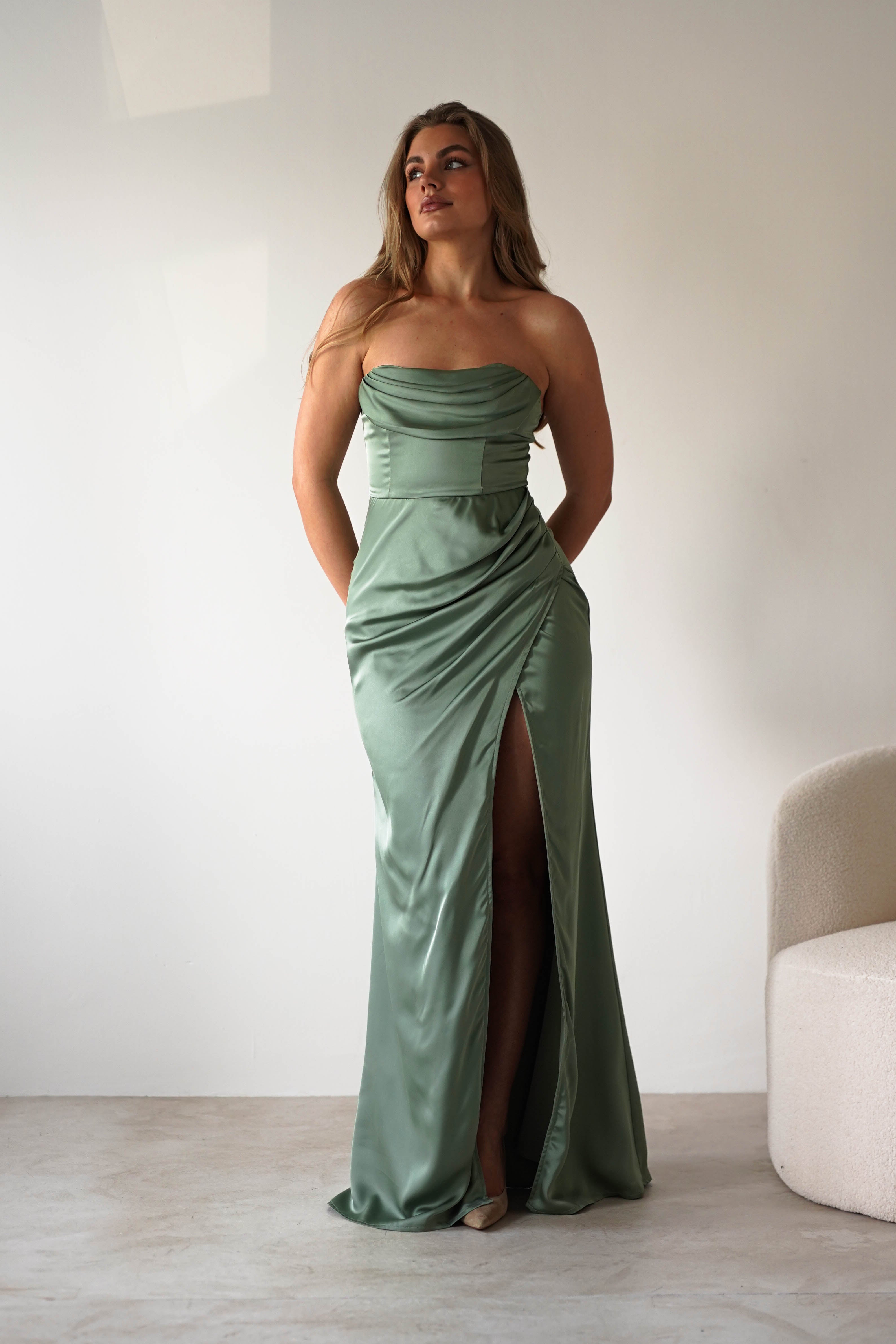 A woman wearing a strapless satin gown in sage green with a high slit stands indoors against a plain white wall, her hands in the dress pockets, looking slightly upward and to the side—perfect for a formal event.