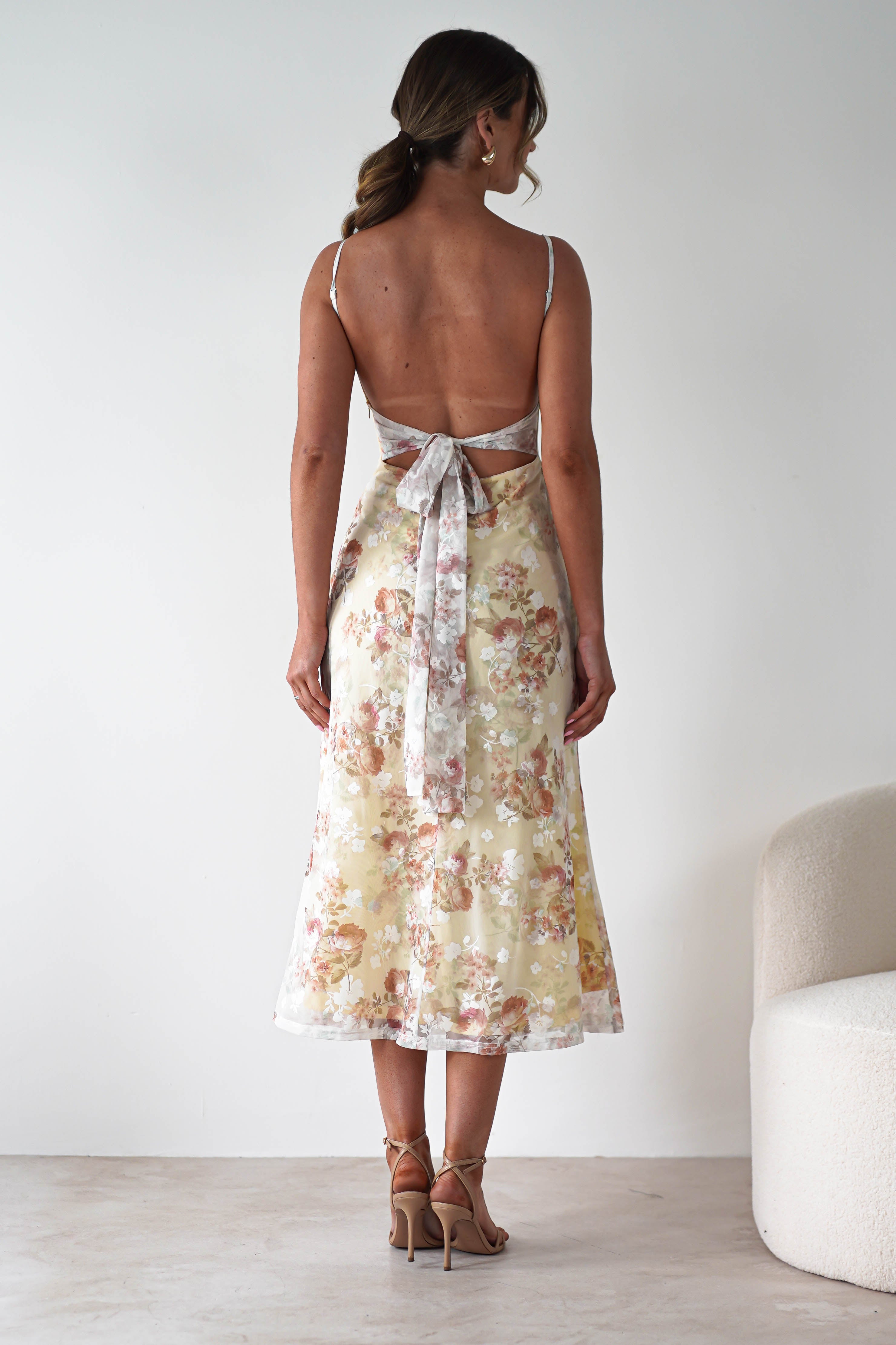 A woman stands indoors with her back to the camera, wearing a floral midaxi dress with spaghetti straps and a bow tied at the back—an elegant choice for a wedding guest. Her light brown hair is in a loose ponytail, paired with nude high-heeled sandals.