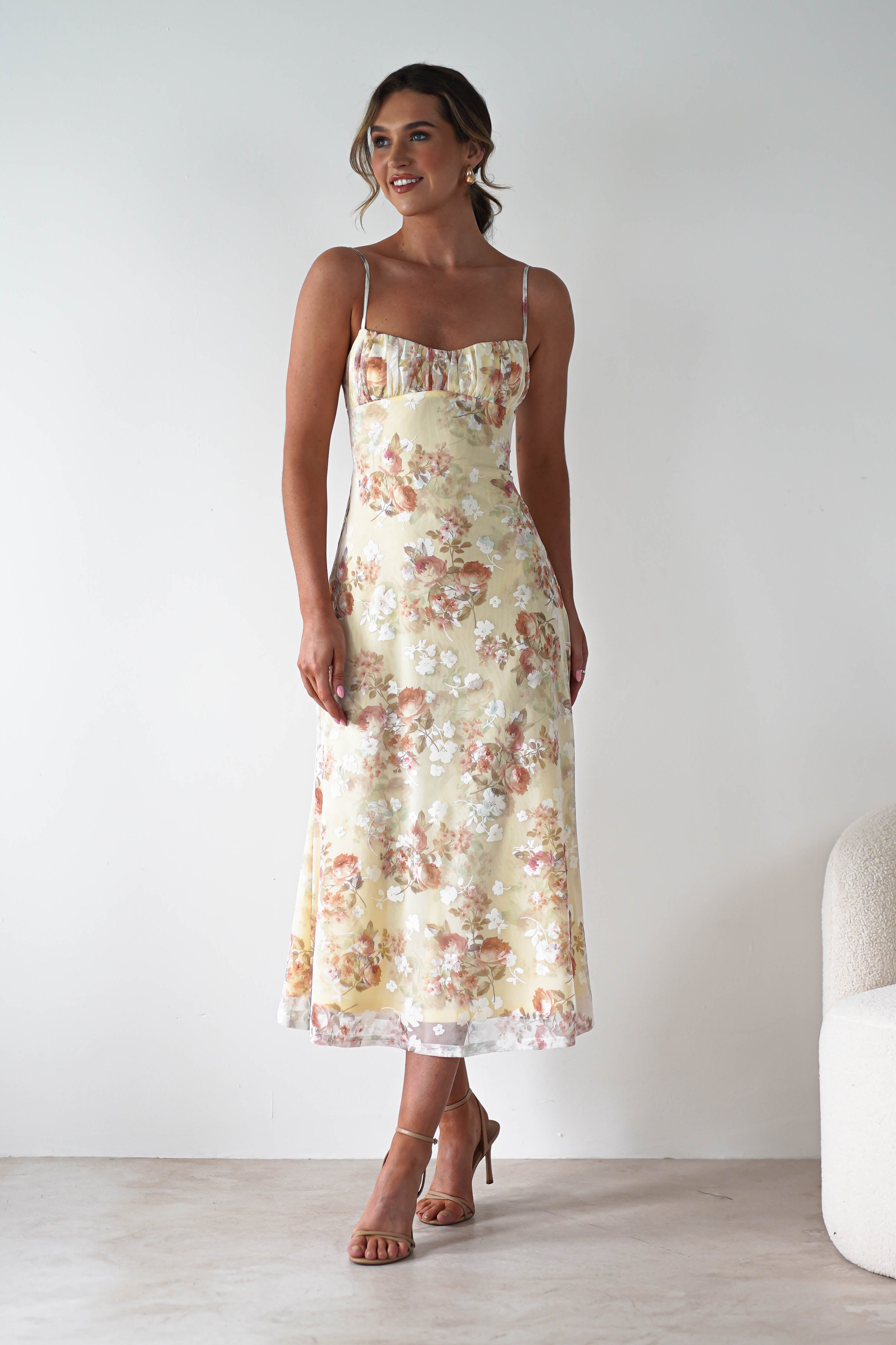 A woman in a light yellow, floral midaxi dress with thin straps and an open back stands indoors, smiling, with her hair in a loose updo. She wears tan strappy heels and poses near a white armchair against a plain background—perfect for a wedding guest.