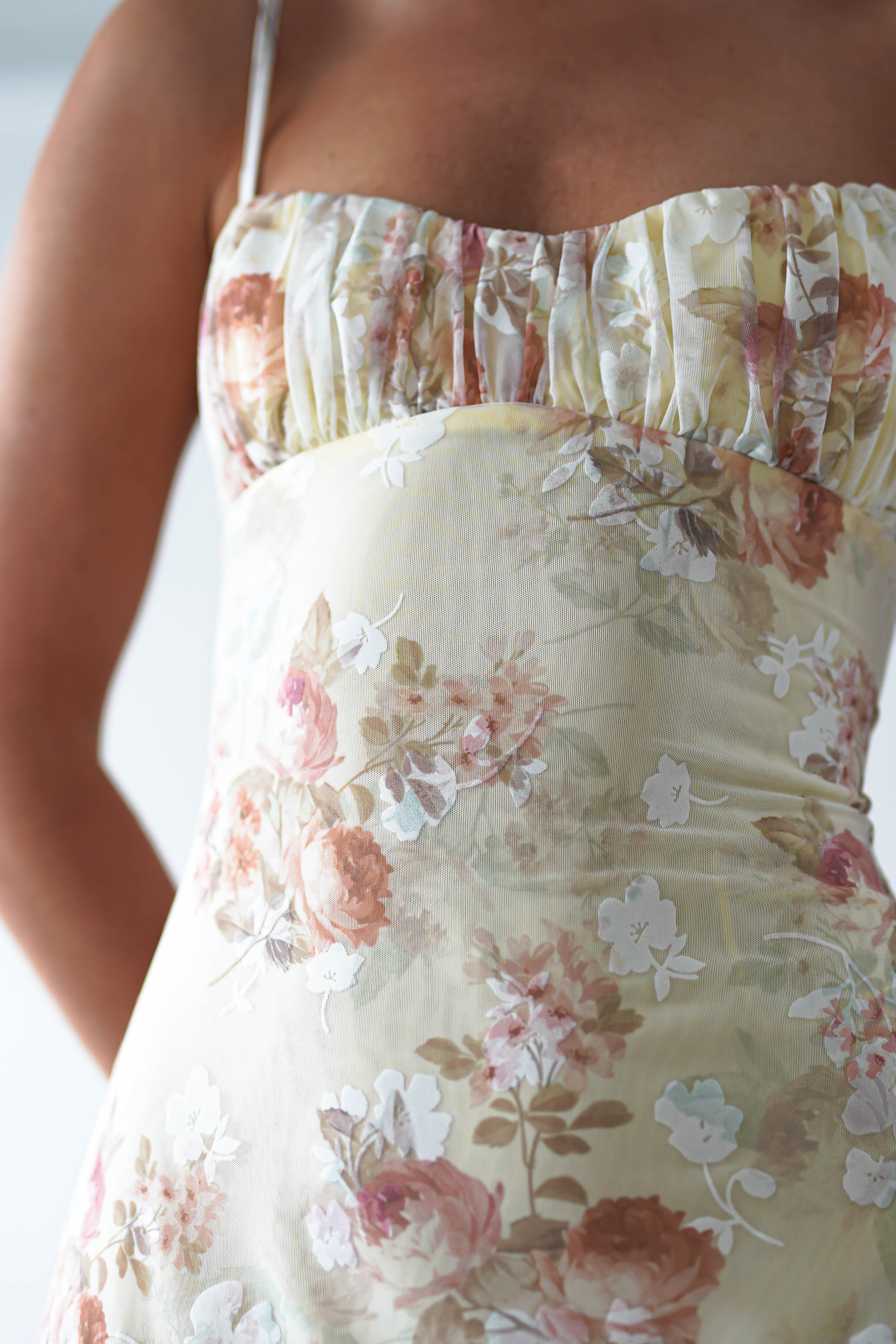 A close-up of a person wearing a sleeveless, floral midaxi dress with gathered fabric at the bust. Featuring soft pastel colors and a pattern of roses and other flowers, its perfect for a wedding guest. The background is blurred and light.