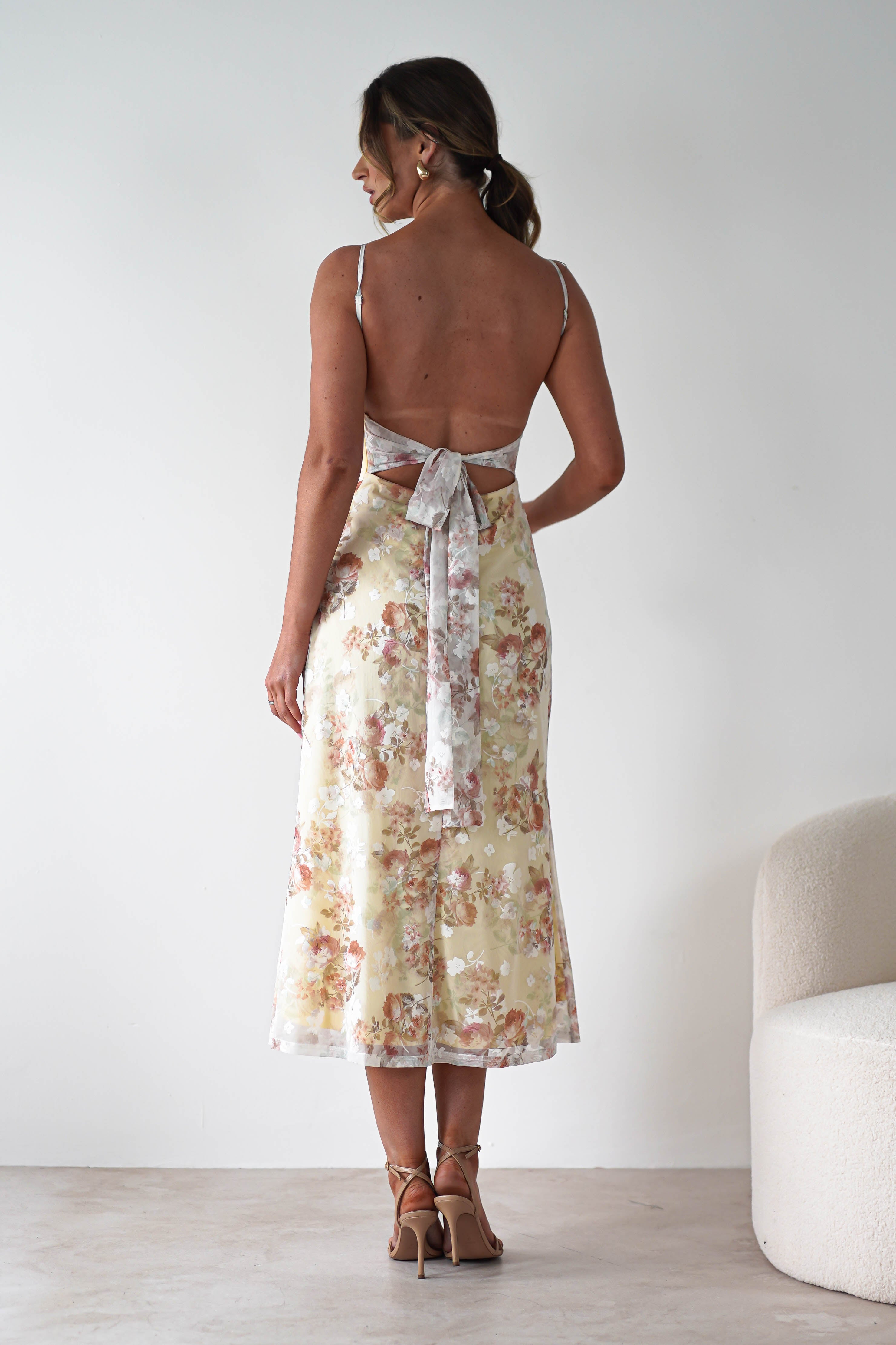 A woman stands facing away, wearing a light yellow floral midaxi dress with an open back and a large bow tied at the waist—perfect for a stylish wedding guest. Her brown hair is in a ponytail, beige heels complete the look, and the background is minimal and neutral.