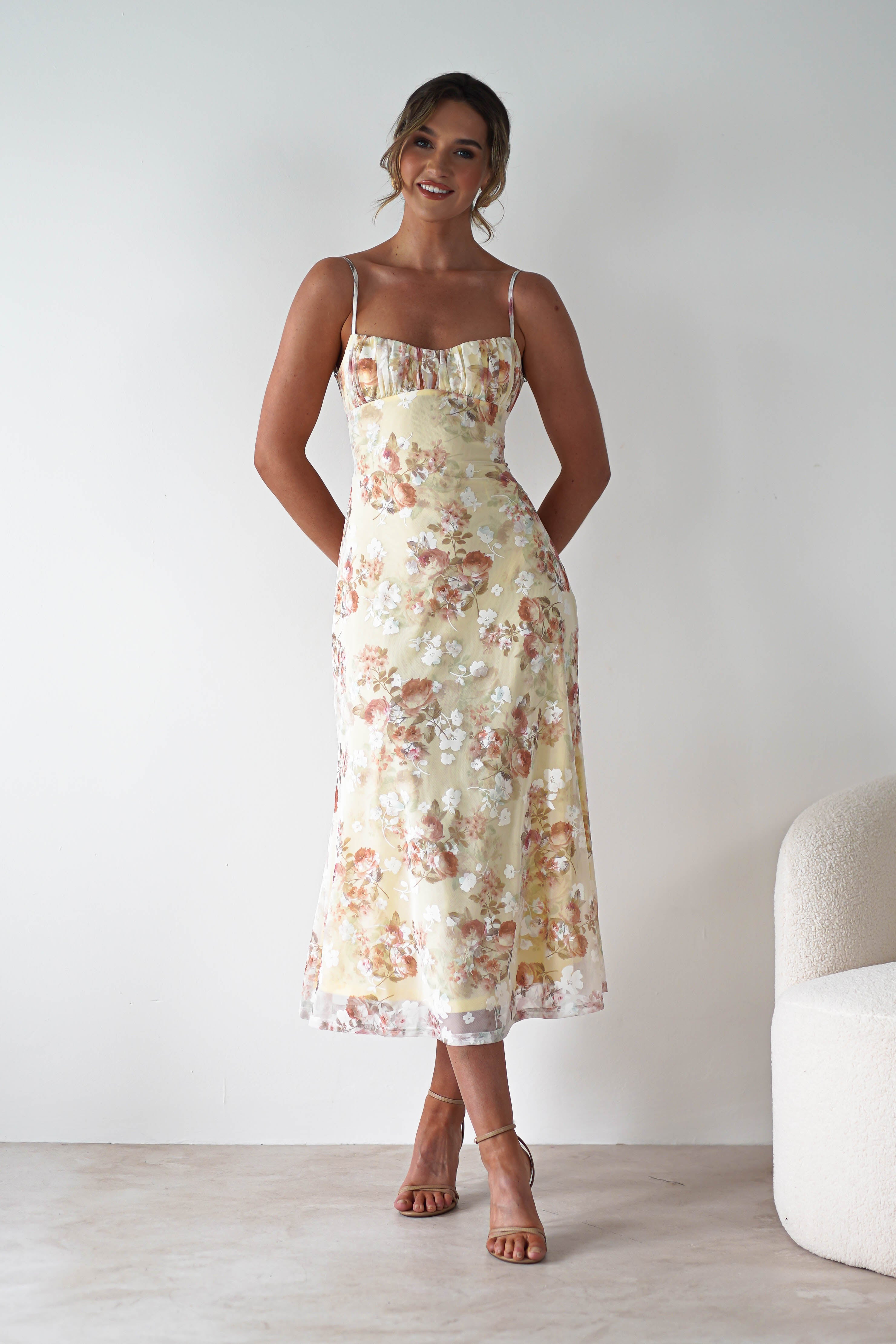 A woman stands smiling against a plain light wall, wearing a light yellow floral midaxi dress with spaghetti straps and tan heels—an elegant look for a wedding guest. Her hair is styled in a loose updo, hands behind her back, with a white textured chair beside her.