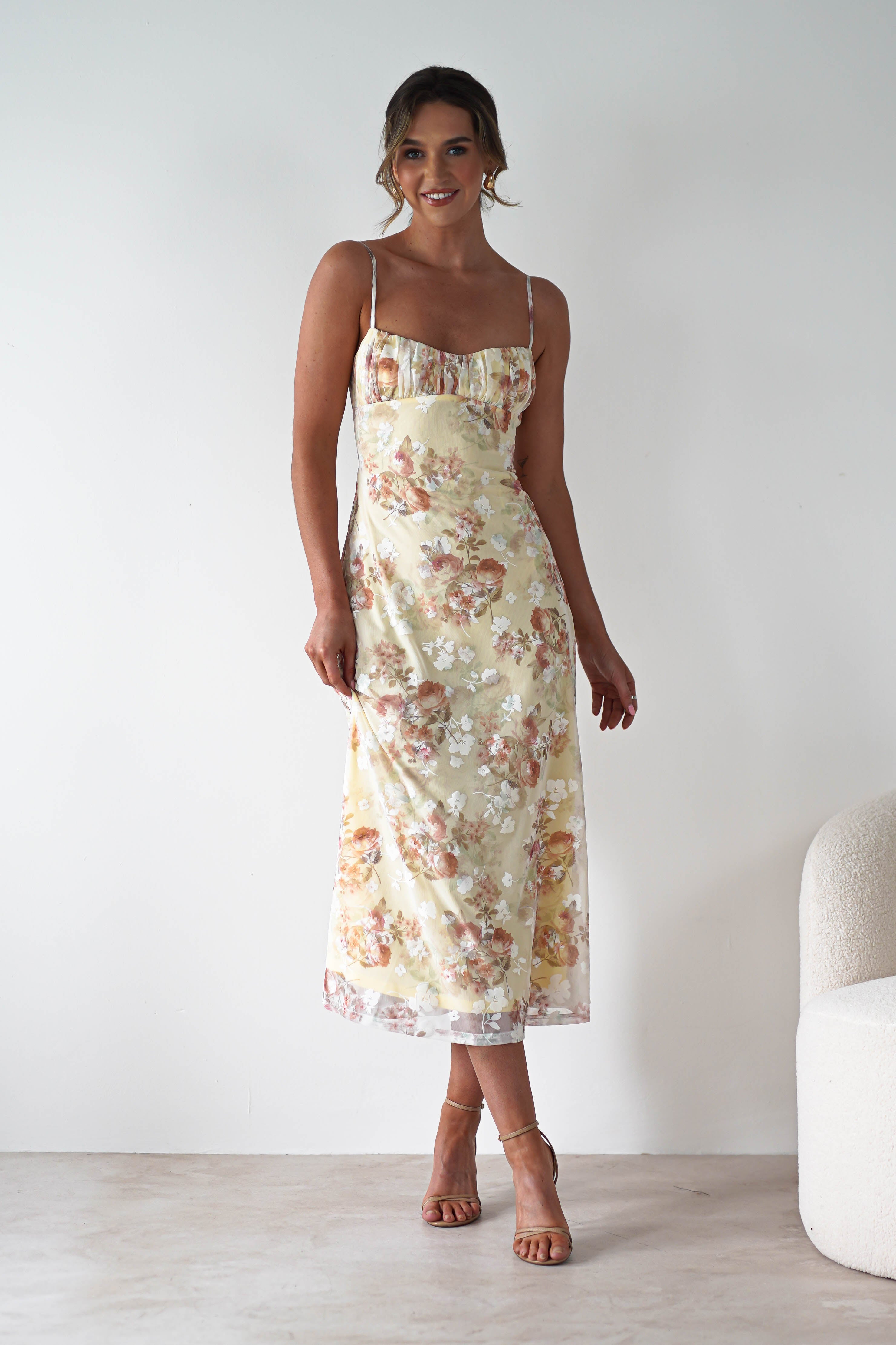 A woman in a sleeveless, light yellow floral midaxi dress with an open back and strappy heels stands smiling against a plain white wall next to a cream-colored chair—perfect for a stylish wedding guest look.