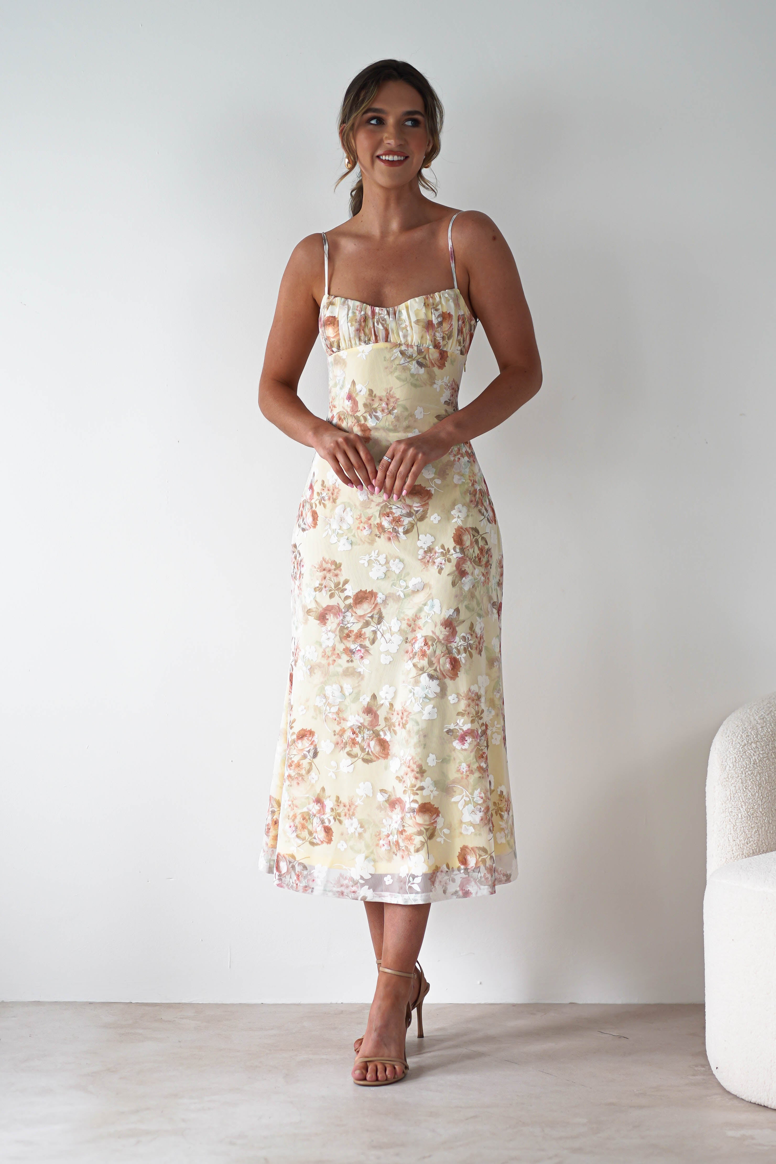 A woman stands indoors against a plain white wall, wearing a light yellow floral midaxi dress with an open back and thin straps—perfect for a wedding guest. She is smiling, with her hands clasped in front of her and nude heels on.