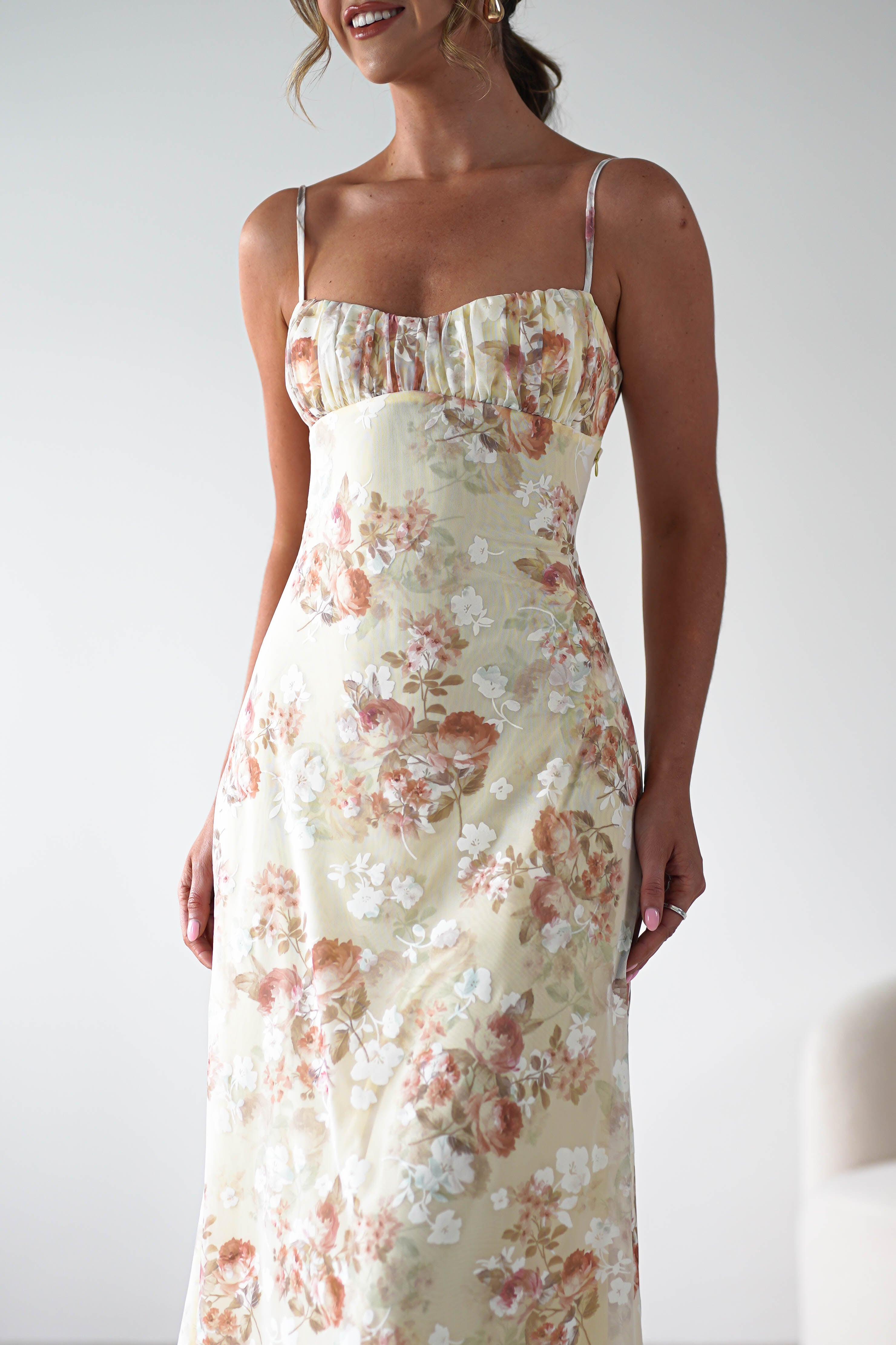 A woman wearing a light yellow, floral midaxi dress with spaghetti straps stands against a plain white background, smiling—perfect for a wedding guest look.
