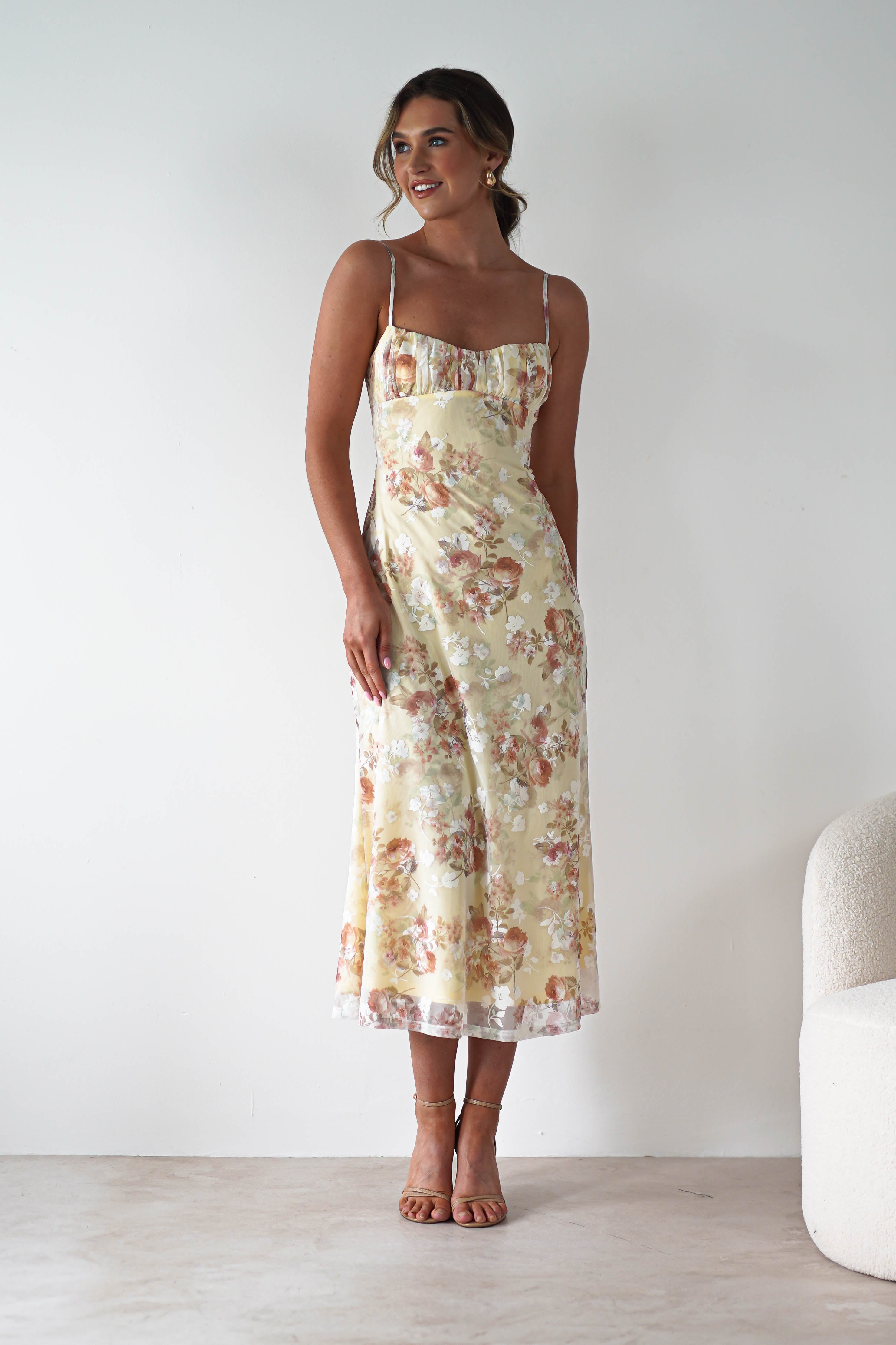 A woman wearing a light yellow floral midaxi dress with spaghetti straps stands indoors, smiling and looking to the side. She has styled hair and strappy heels—an ideal look for a wedding guest. A cream-colored chair is partially visible beside her.