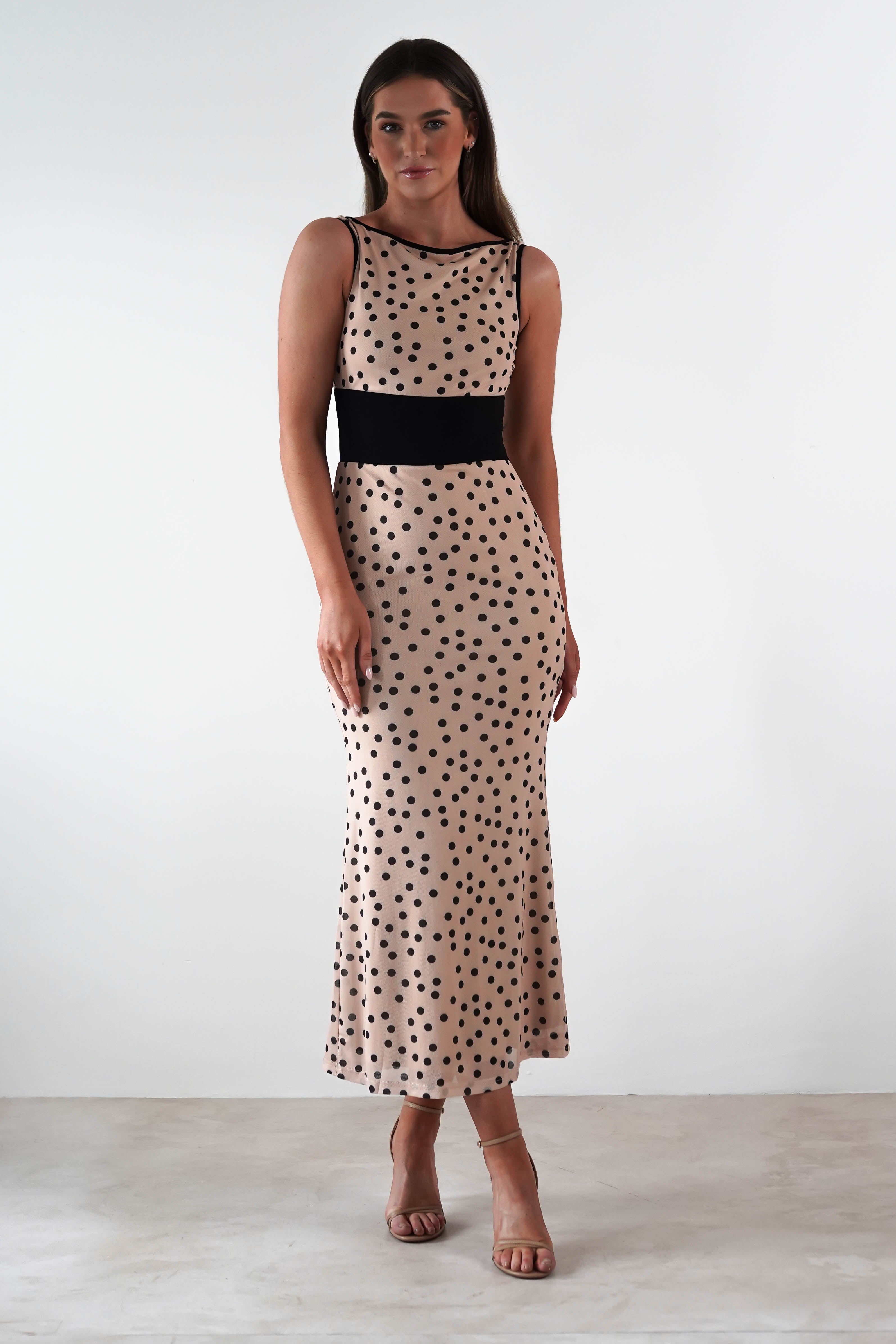 In a neutral-colored room, a woman stands wearing a beige wedding guest dress adorned with black polka dots and a classy black waistband. Her long brown hair complements the high-heeled sandals, and her calm expression gazes directly at the camera.