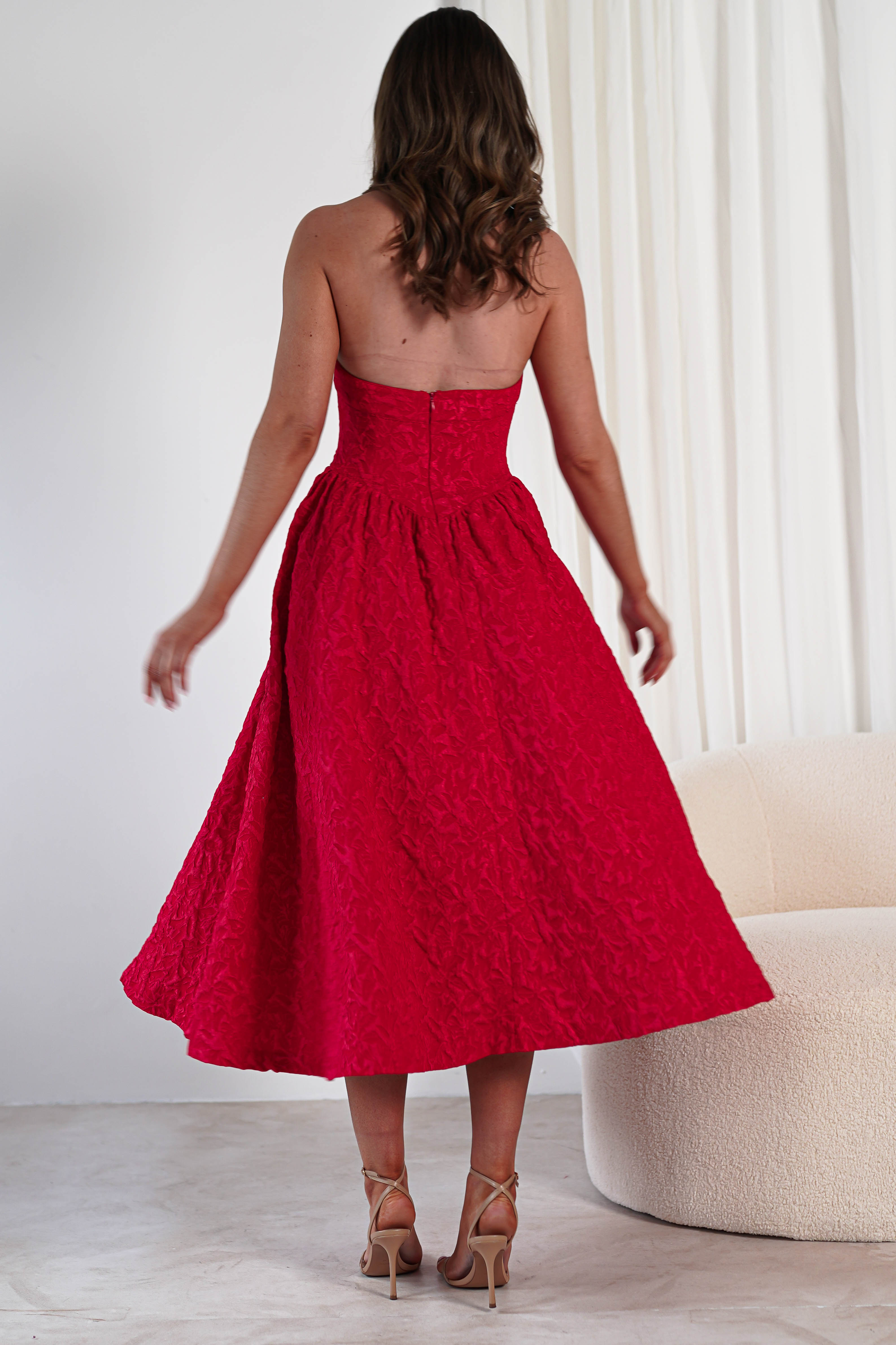 A woman with wavy brown hair wearing a strapless, textured red midi dress with pockets and beige high heels stands facing away in a modern, minimal room with white curtains and a cream-colored chair.