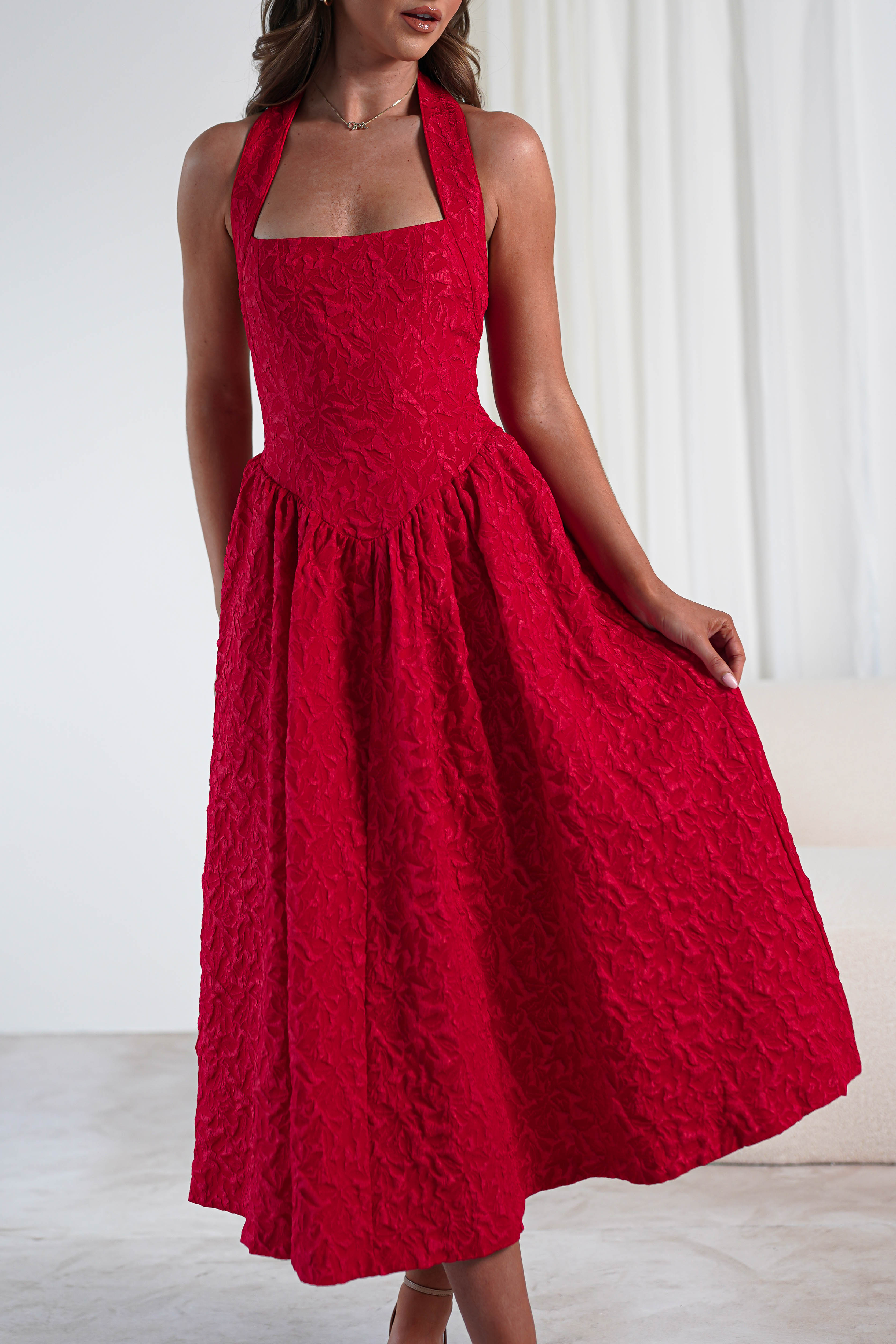A woman wearing a sleeveless, textured red halter dress with a fitted bodice and flared skirt stands indoors, holding out one side of her skirt with pockets. The background features white curtains and light-colored furniture.