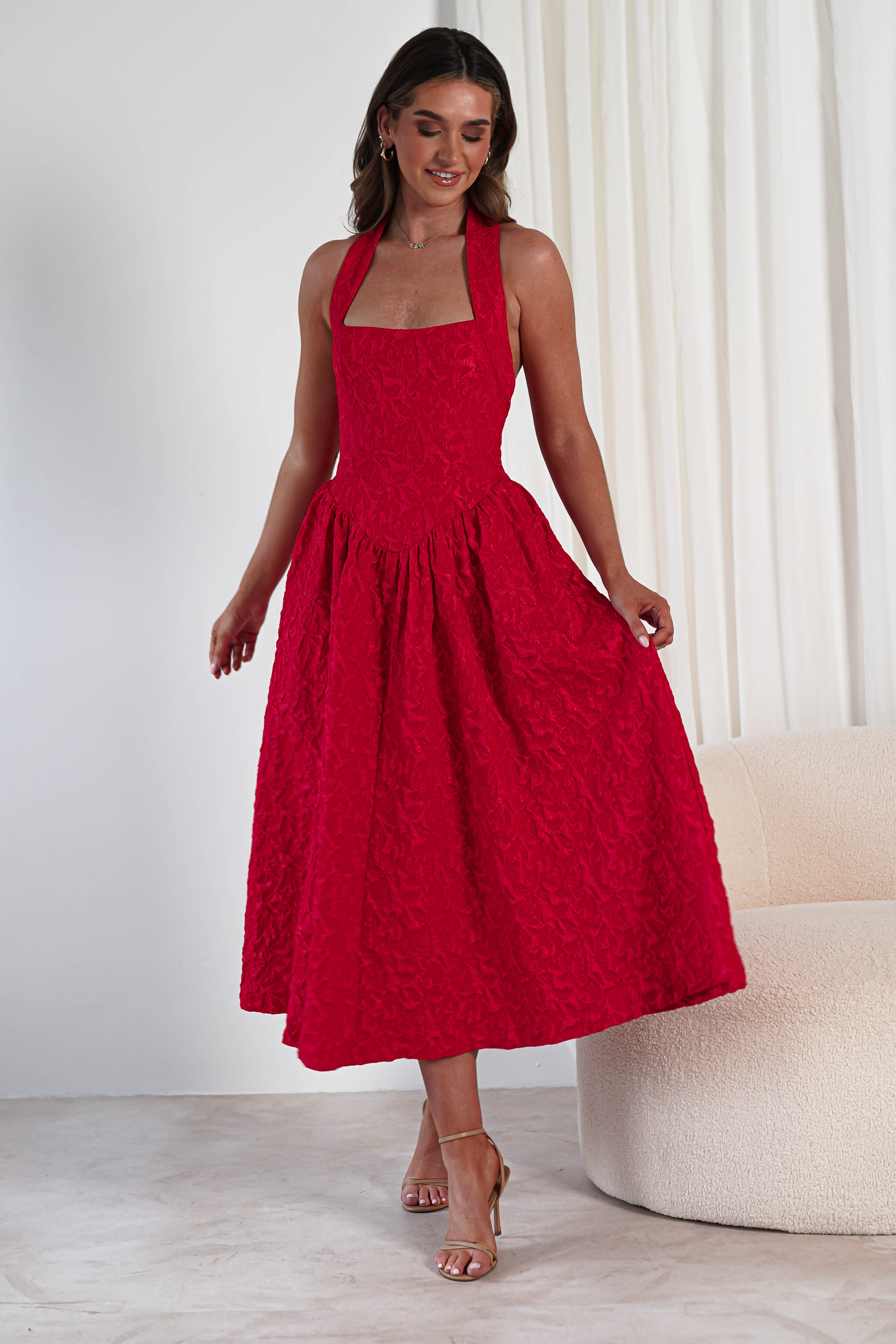 A woman stands indoors wearing a sleeveless, textured fabric red midi dress with a square neckline and halter straps. She holds the skirt out on both sides, revealing discreet pockets, posing in front of white curtains and a cream sofa.