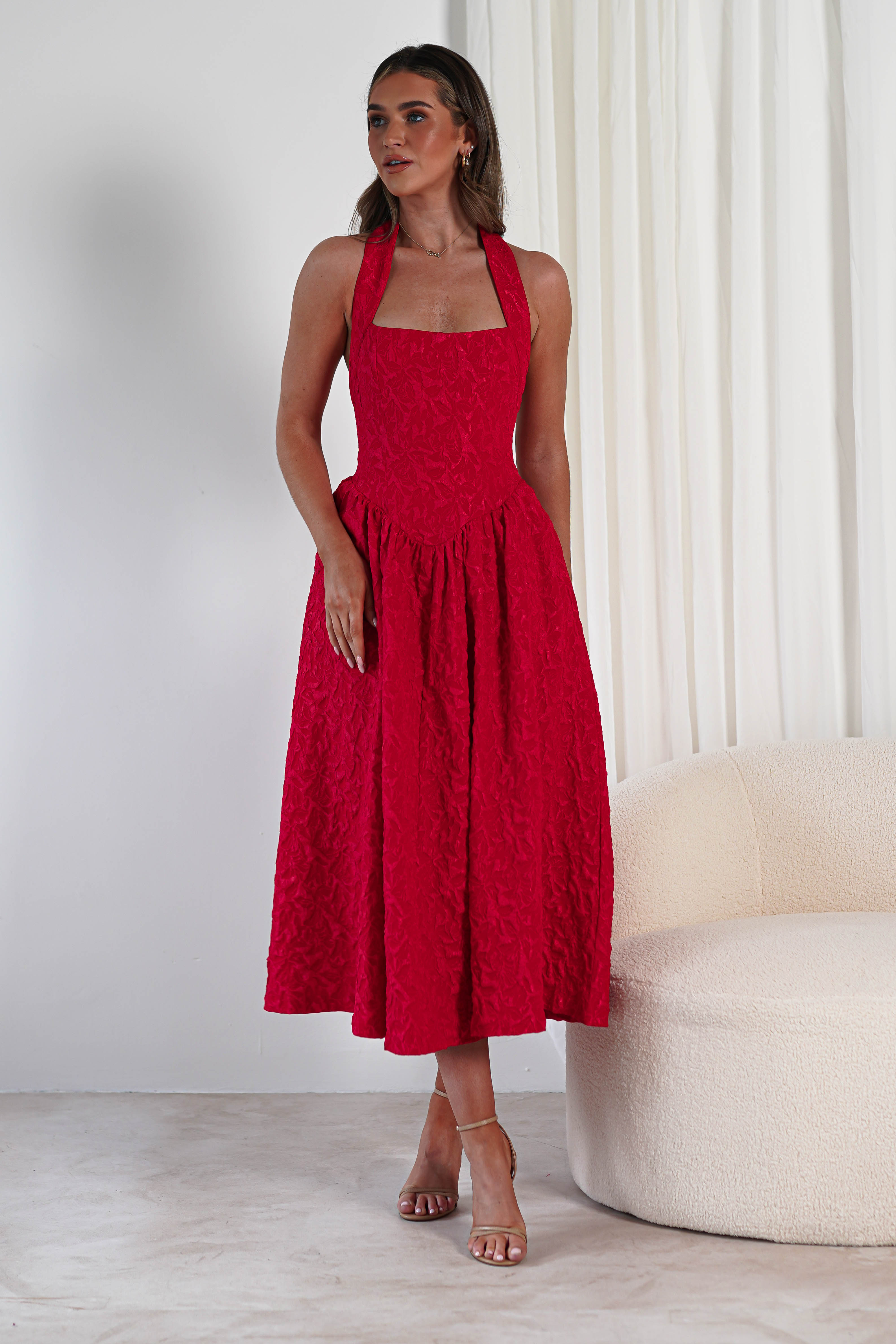 A woman stands indoors in a bright red, textured fabric halter-neck midi dress with a fitted bodice, flared skirt, and pockets—perfect for formal events. She has light brown hair, strappy nude heels, and is framed by white curtains and a cream sofa.