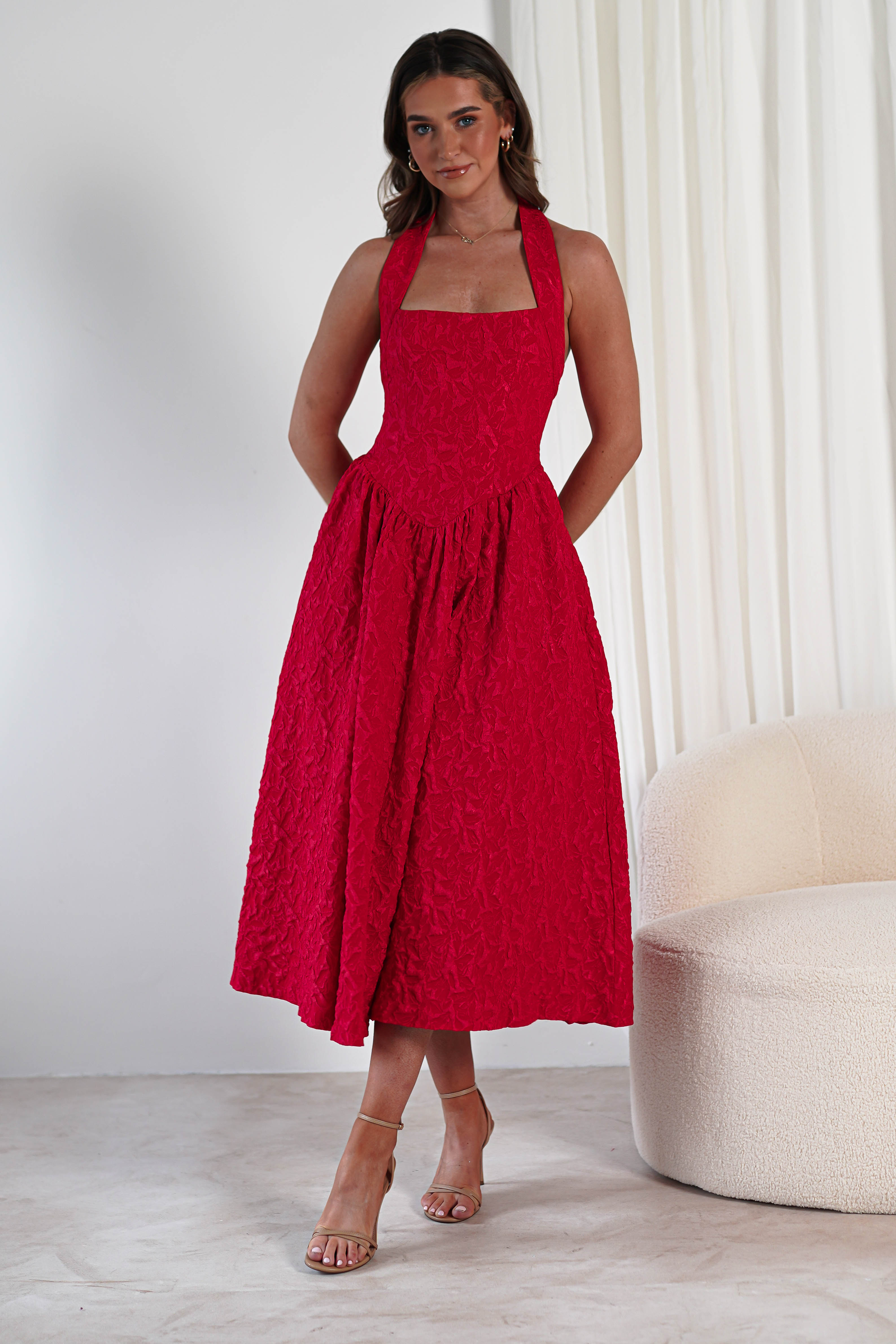 A woman stands indoors in a bright red, textured fabric midi dress with a halter neckline and hidden pockets—perfect for formal events. She poses with her hands behind her back, nude heeled sandals on, beside a cream-colored chair.