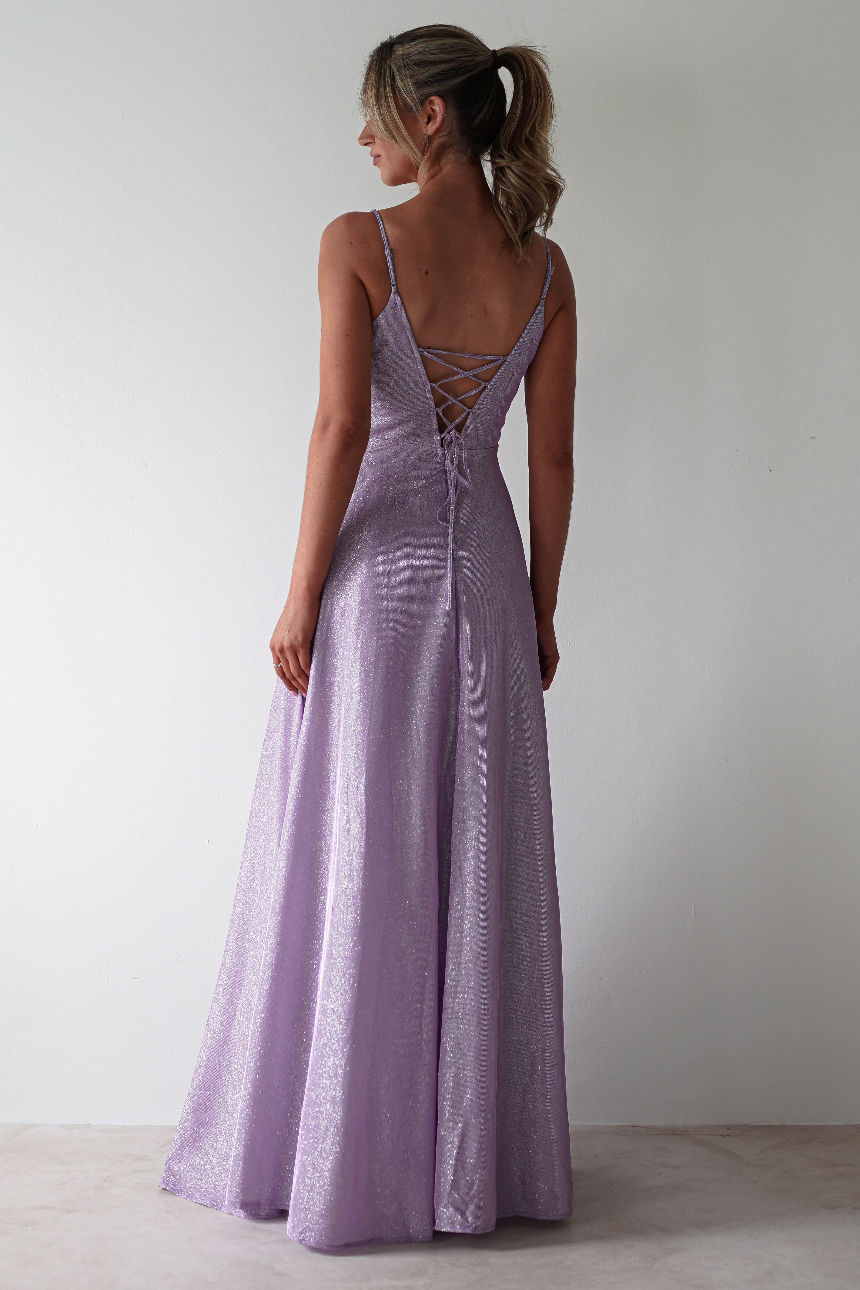 A woman in a lavender, glitter gown stands with her back to the camera. The dress features diamante straps and a lace-up back. Her hair is in a ponytail, perfectly styled for formal events, as she poses against a plain, light-colored background.