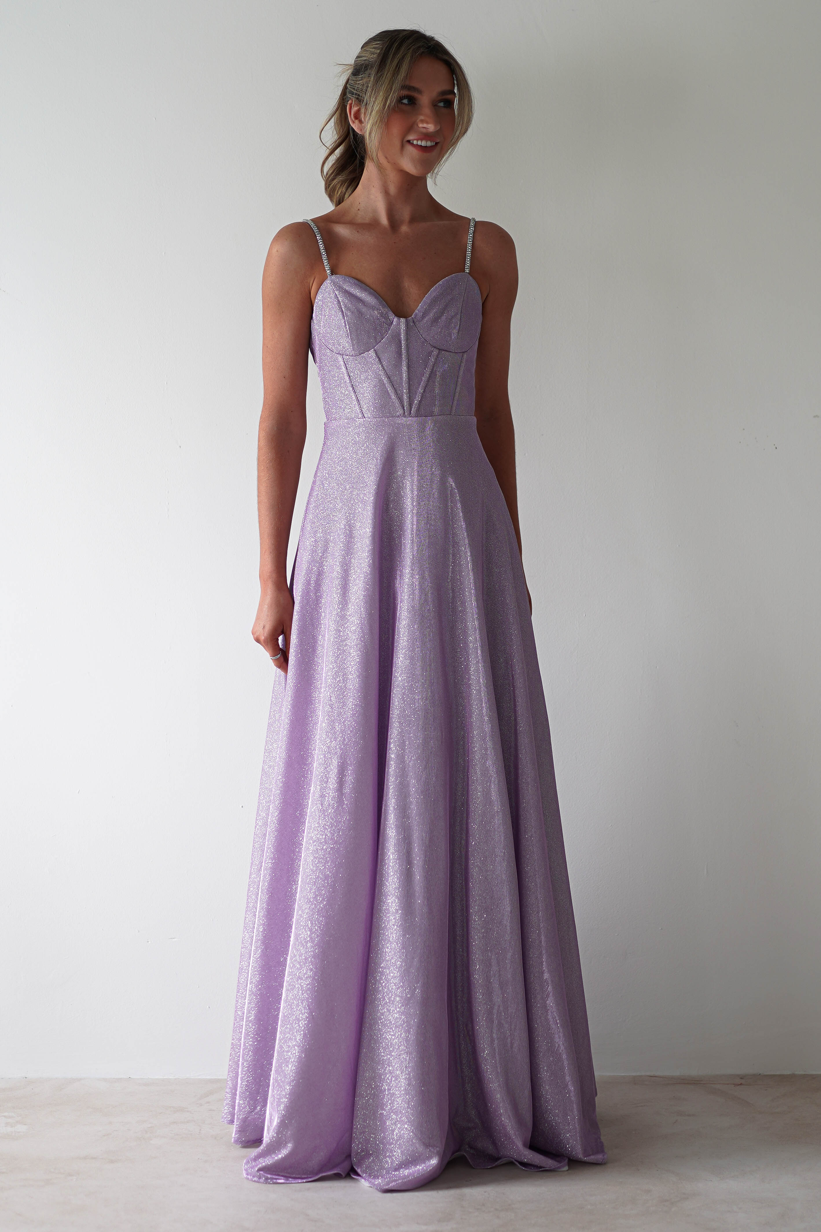 A woman is wearing a long, glittering lavender gown with thin diamante straps and a fitted bodice. She stands against a plain white background, smiling slightly, with her hair styled in an elegant updo, ready for formal events.