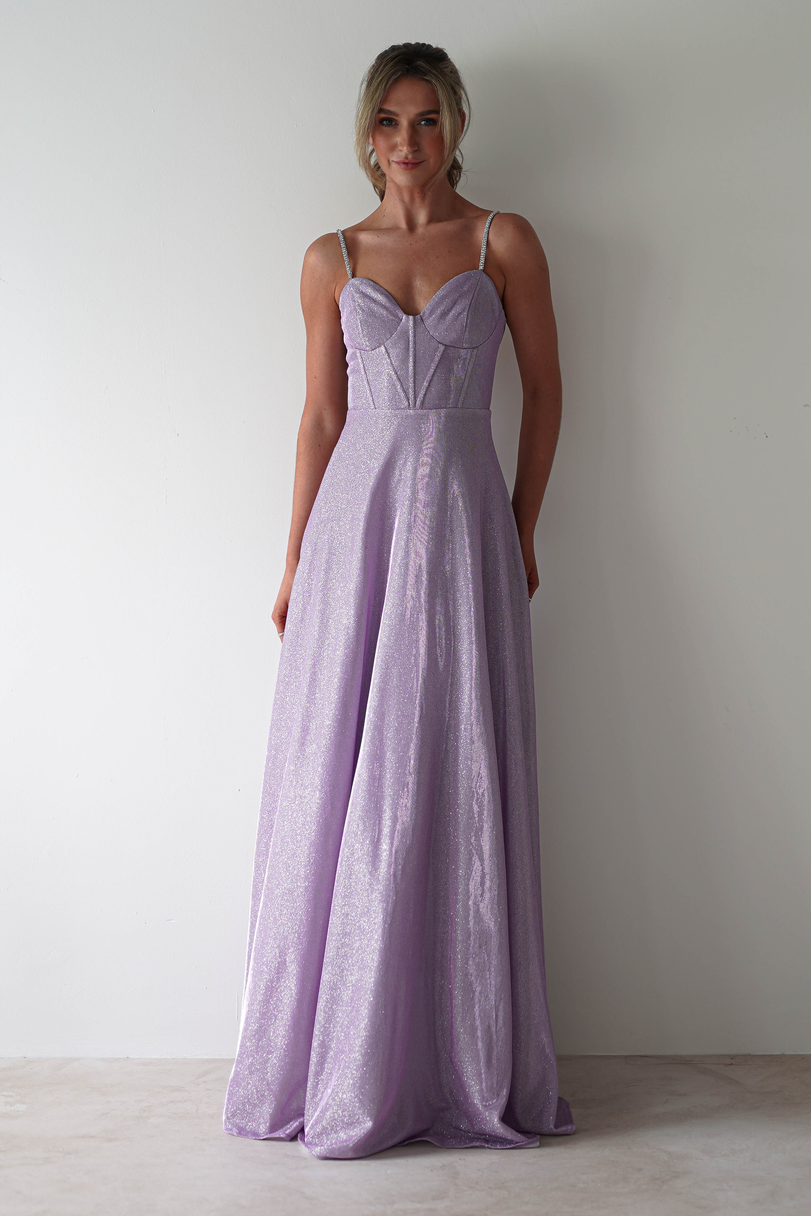 A person dons a light purple, sleeveless floor-length gown with a fitted bodice and subtle diamante straps. They stand against a plain white background, their hair styled in a loose updo—an ideal look for formal events.