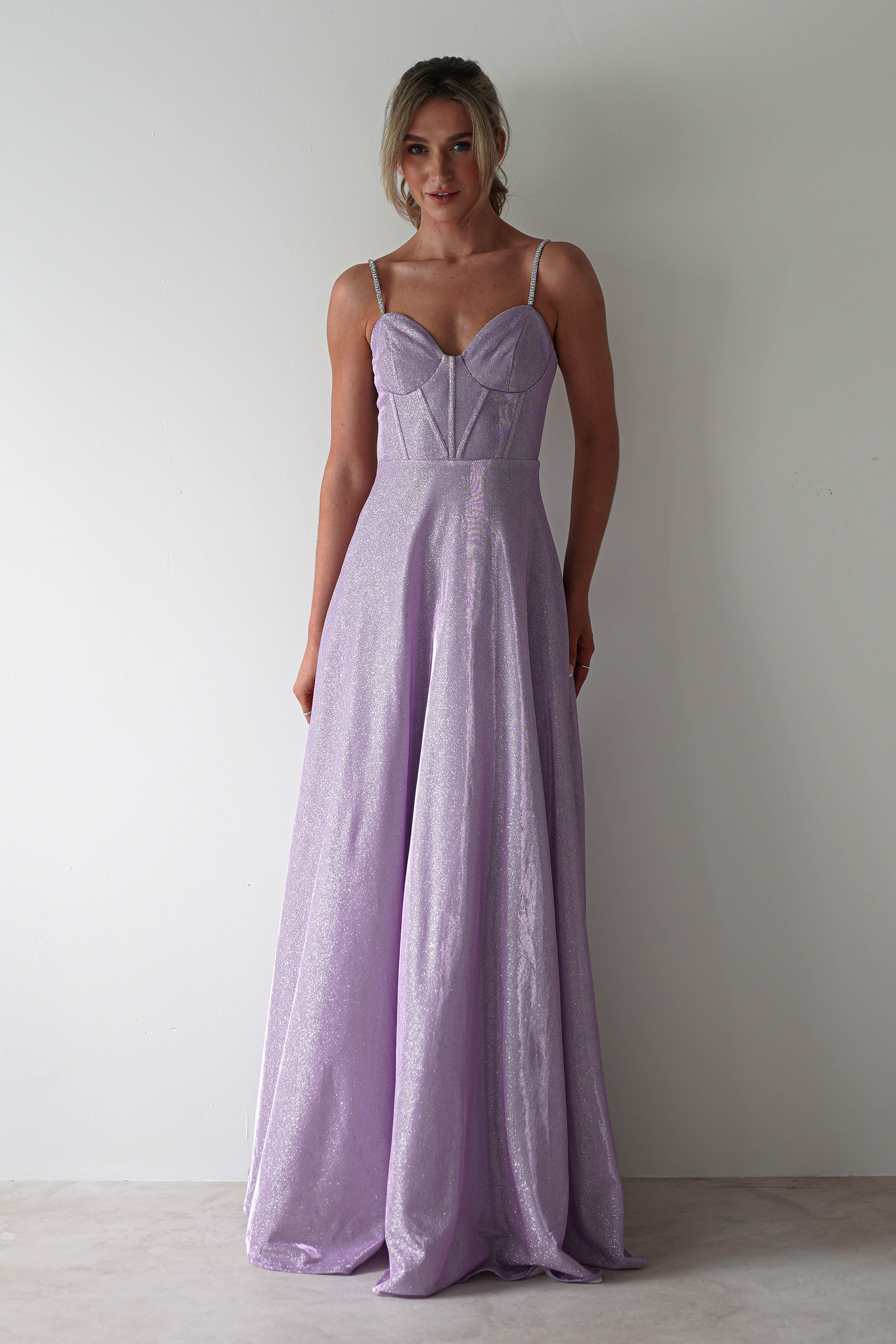 A person in a glitter gown with diamante straps stands against a plain, light-colored background. The shimmering lavender dress features a fitted bodice and a flowing skirt. Their hair is tied up, and they are smiling slightly, ready for formal events.