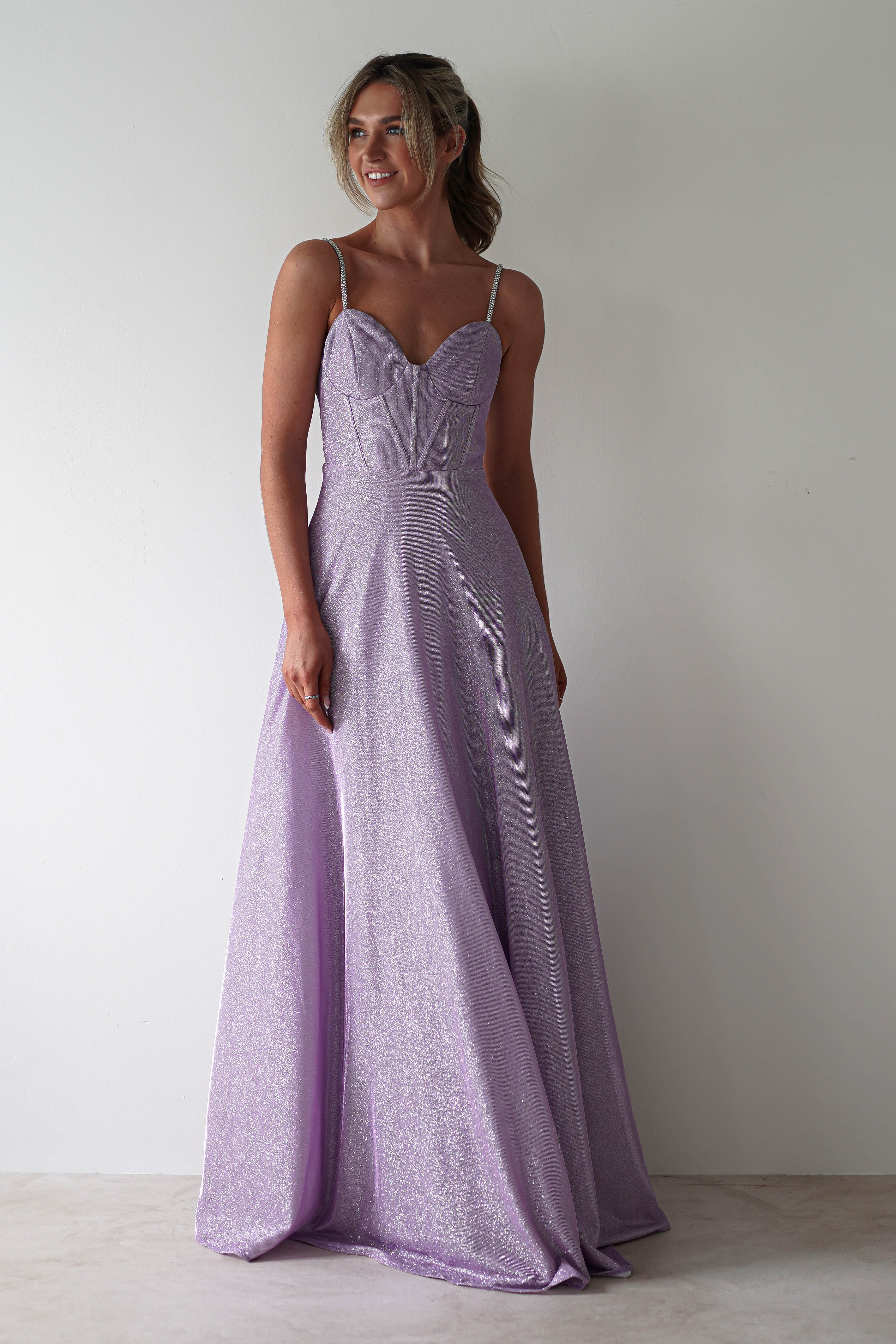 Woman wearing a sparkling lavender ball gown with diamante straps, standing against a plain white background. She has her hair styled up and looks to the side with a soft smile, showcasing the dresss floor-length skirt and fitted bodice—perfect for formal events.