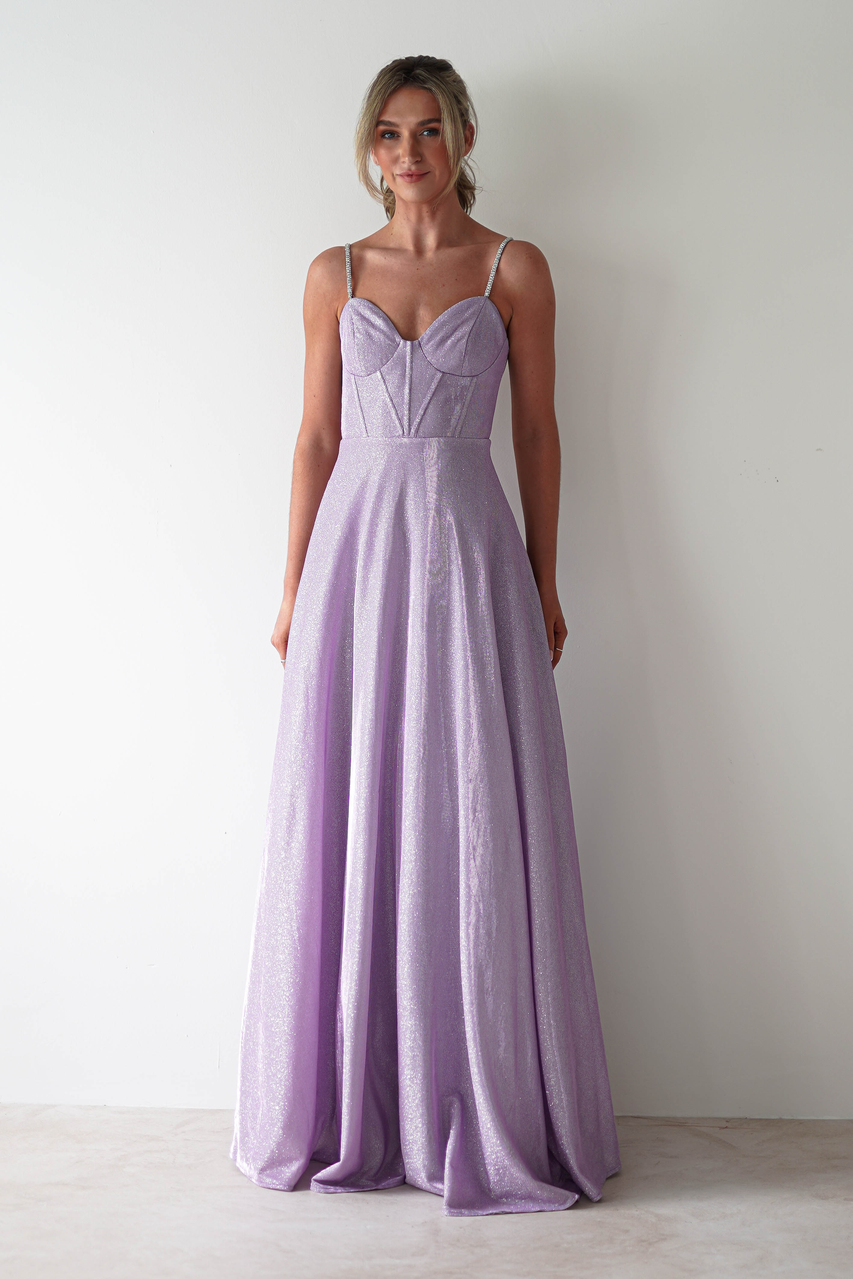 A person stands against a white background, gracefully wearing a sleeveless, glitter gown in shimmering lavender. The dress features diamante straps and a fitted bodice, flowing into a long, full skirt. Their hair is styled up as they look forward with poise.