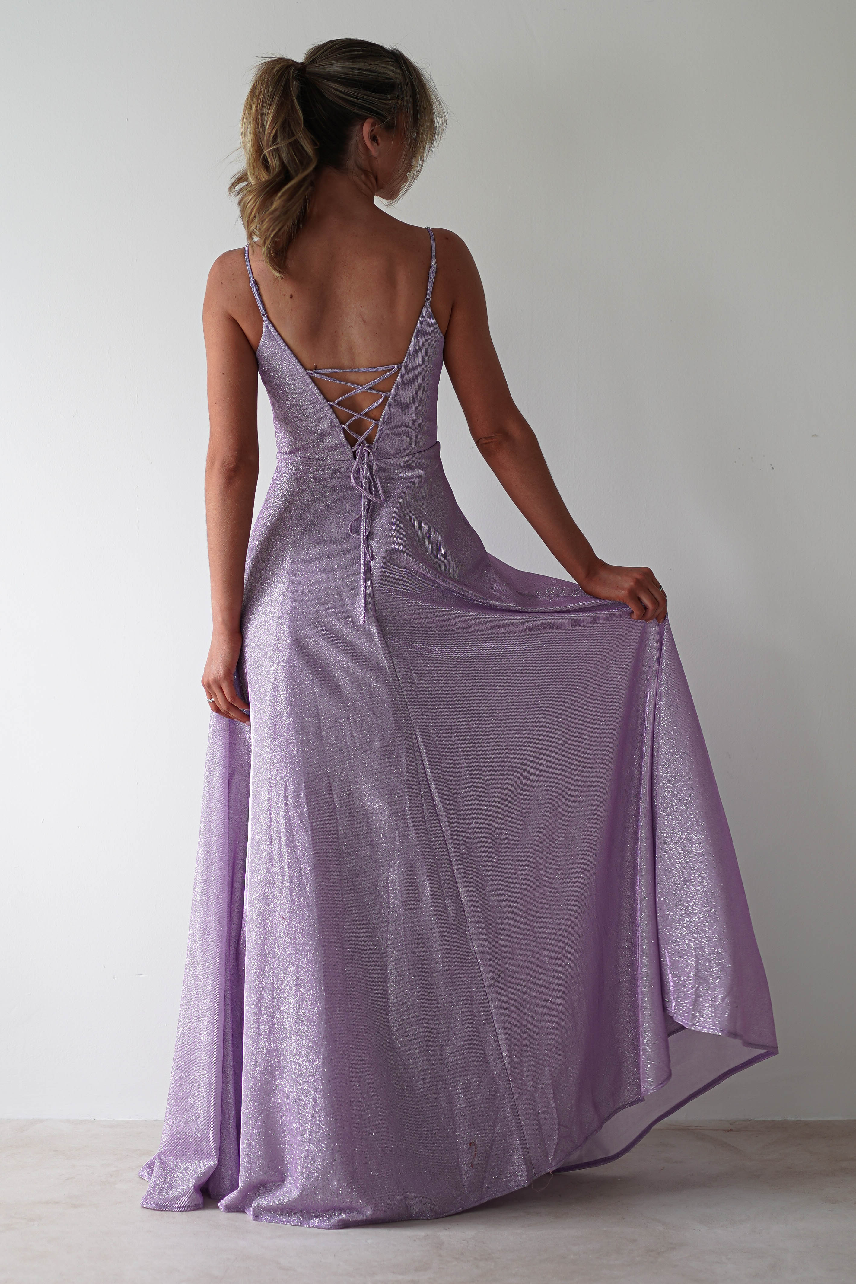 A woman in a glitter gown featuring shimmering lavender fabric and diamante straps stands against a plain white background, gently holding the dresss fabric to the side—perfect for formal events.