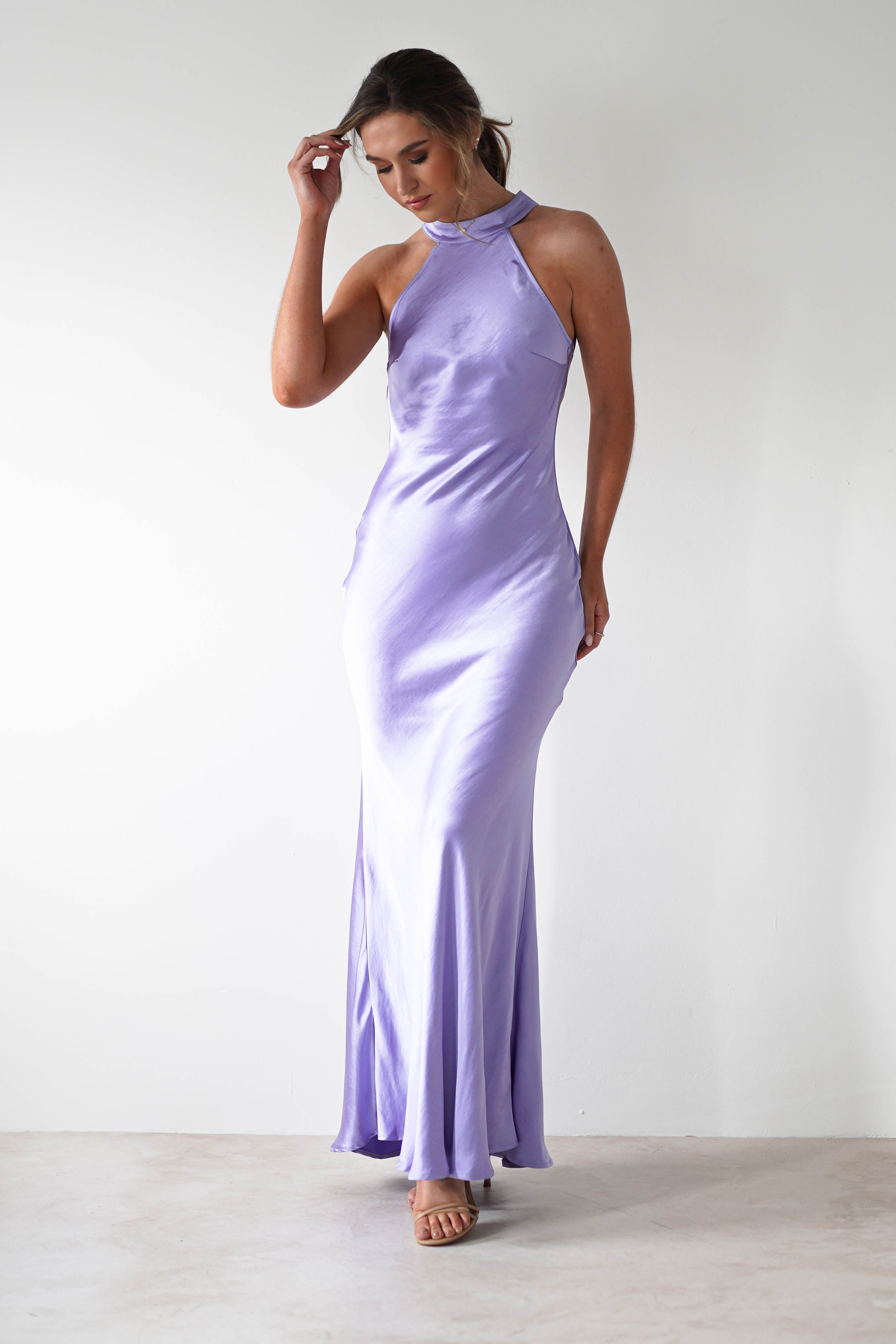 A woman stands against a plain white background wearing a lilac satin material halterneck maxi dress with an open-back detail. She looks down, touching her hair with one hand, and pairs the look with nude high-heeled sandals.