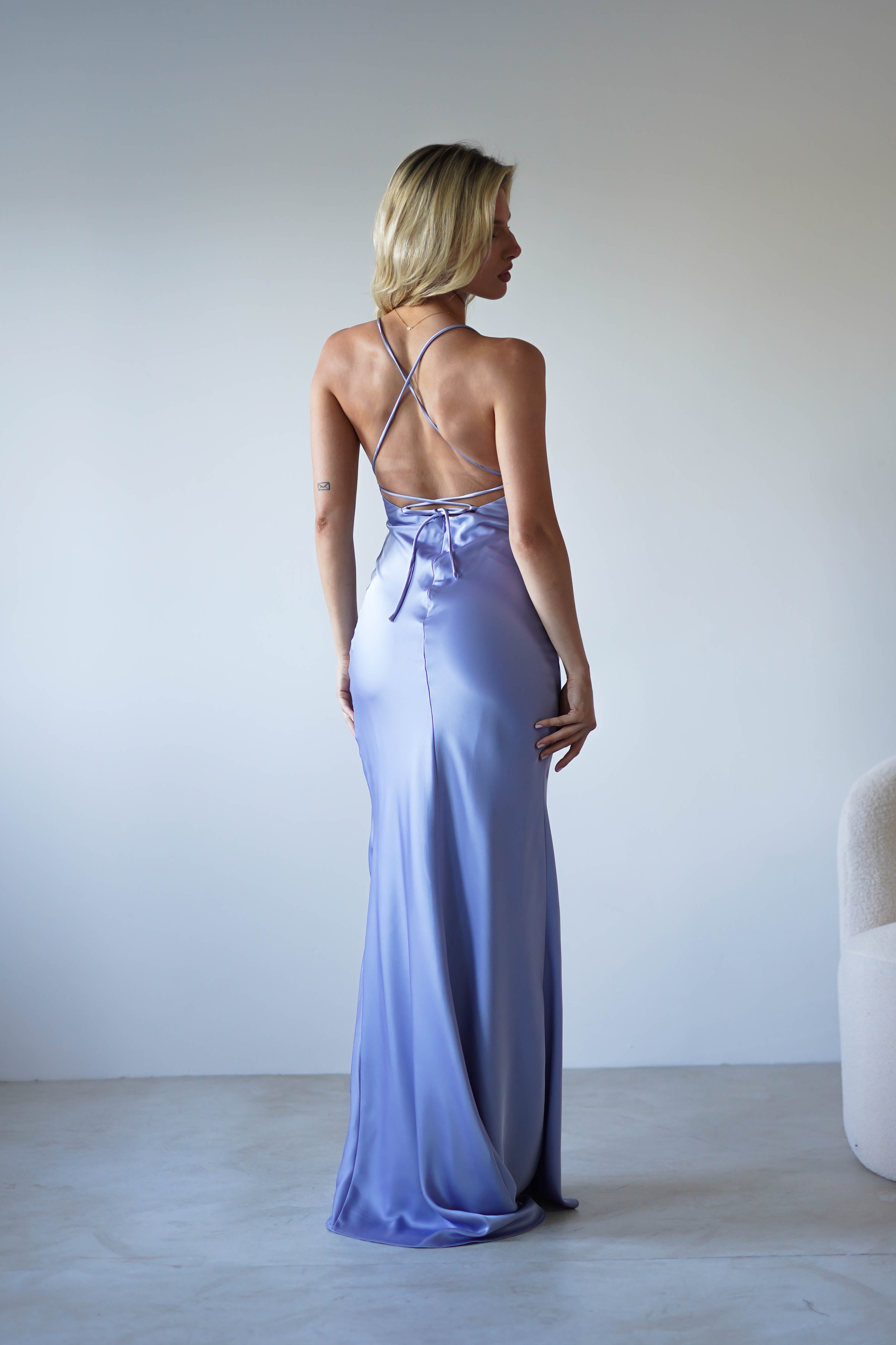A woman with blonde hair stands facing away, wearing a figure slimming, light purple satin dress with thin crisscrossed straps—perfect for formal events. She is in a minimal, modern room with a neutral-toned chair nearby.