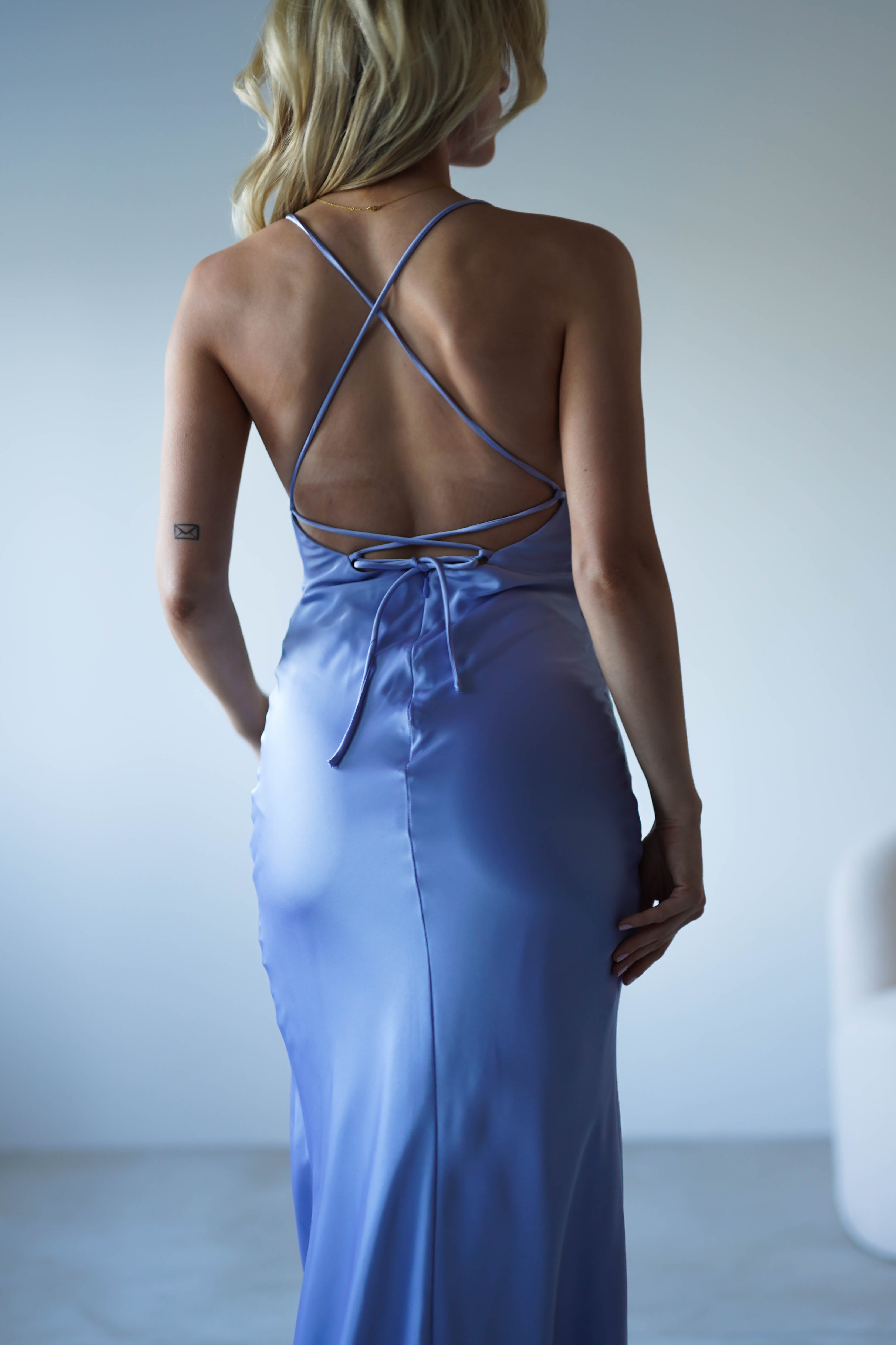 A woman with blonde hair is seen from behind wearing a figure slimming light blue satin dress with crisscross straps and an open back. She stands in a softly lit, minimalistic room, ready for formal events.