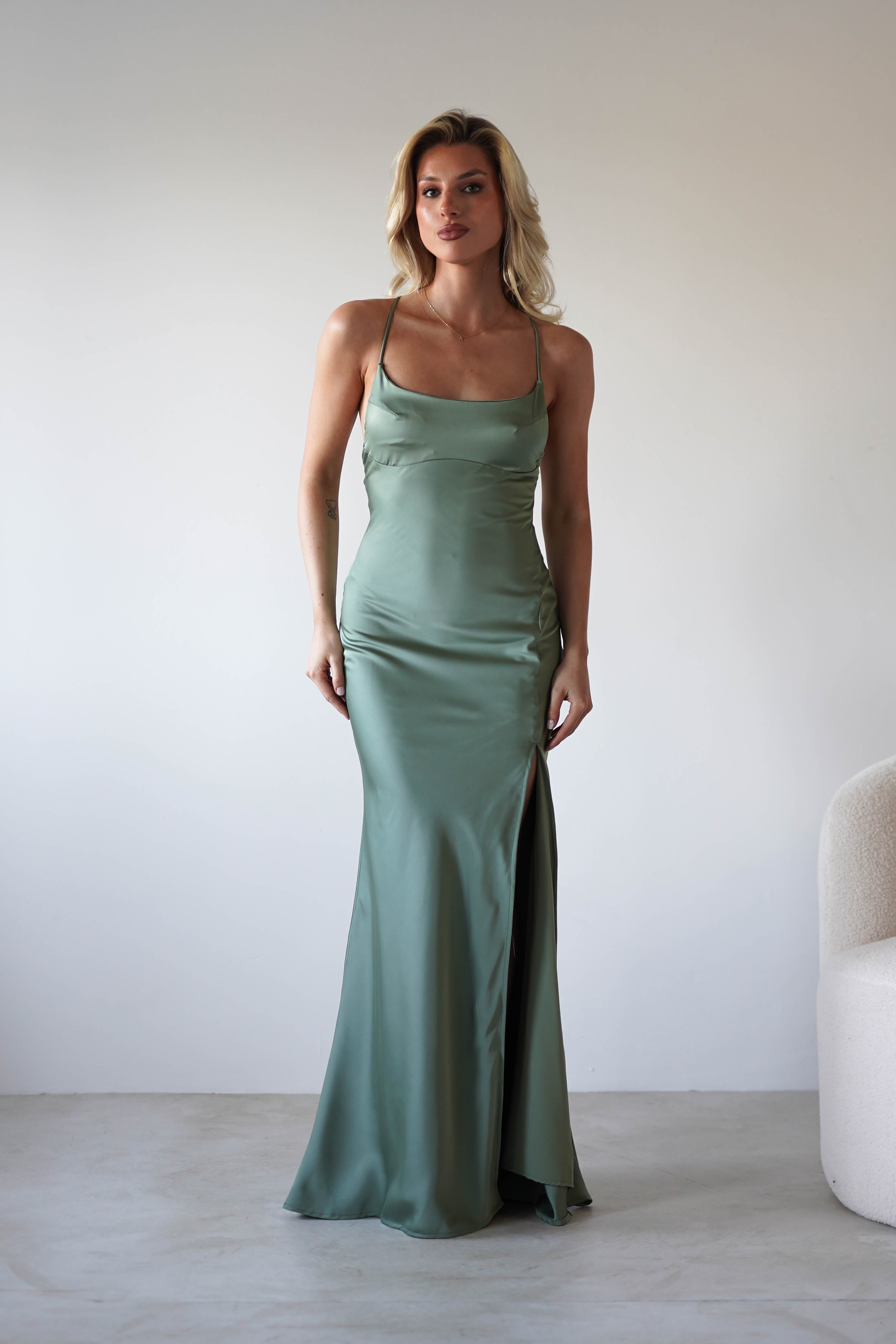 A woman stands indoors wearing a long sage green satin slip dress with adjustable tie straps and a thigh-high slit. She has blonde hair and is posing in front of a plain light-colored wall next to a white armchair.