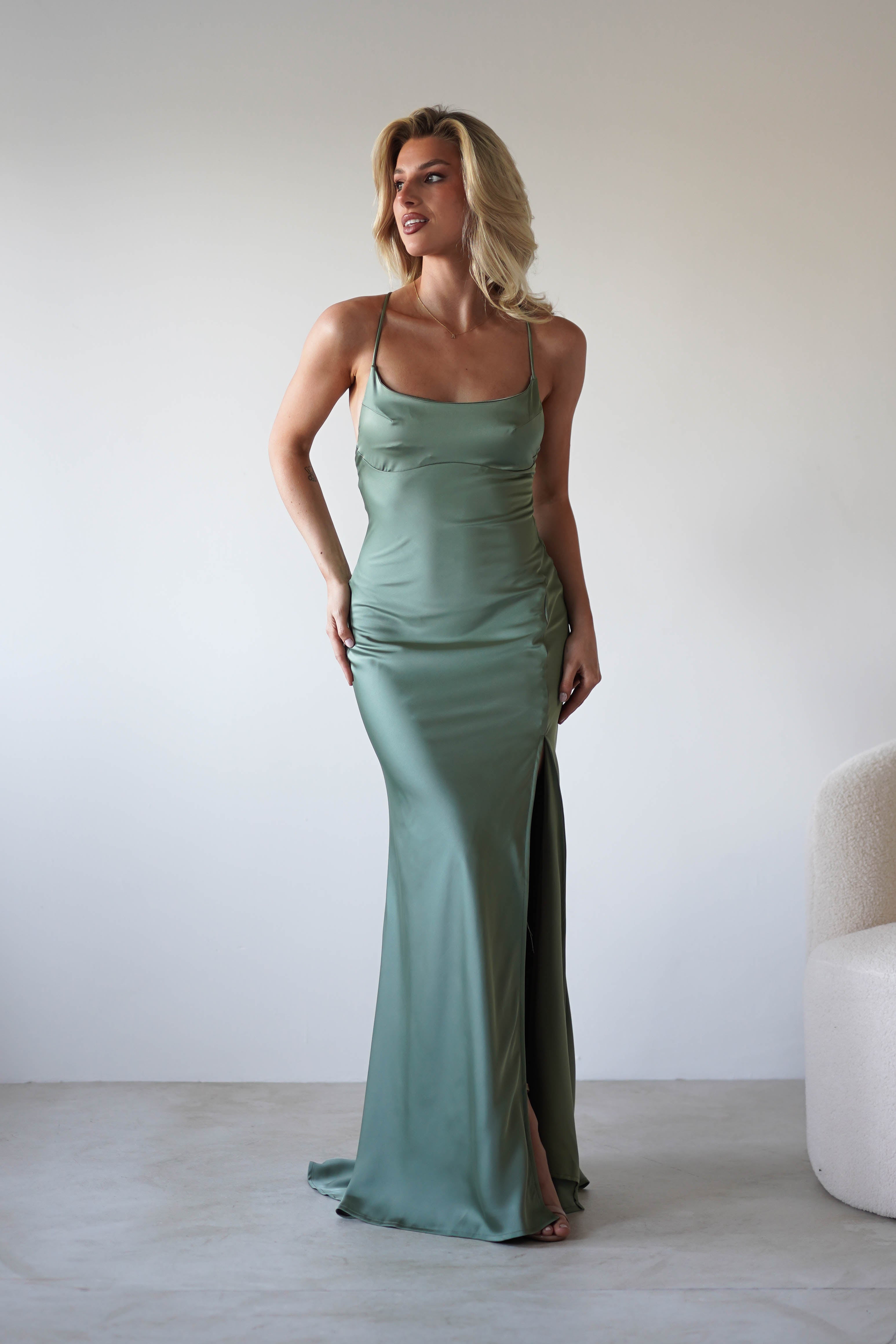 A woman with blonde hair wears a long, sage green satin slip dress with adjustable tie straps and a thigh-high slit. She stands indoors near a white chair, smiling and looking to the side.