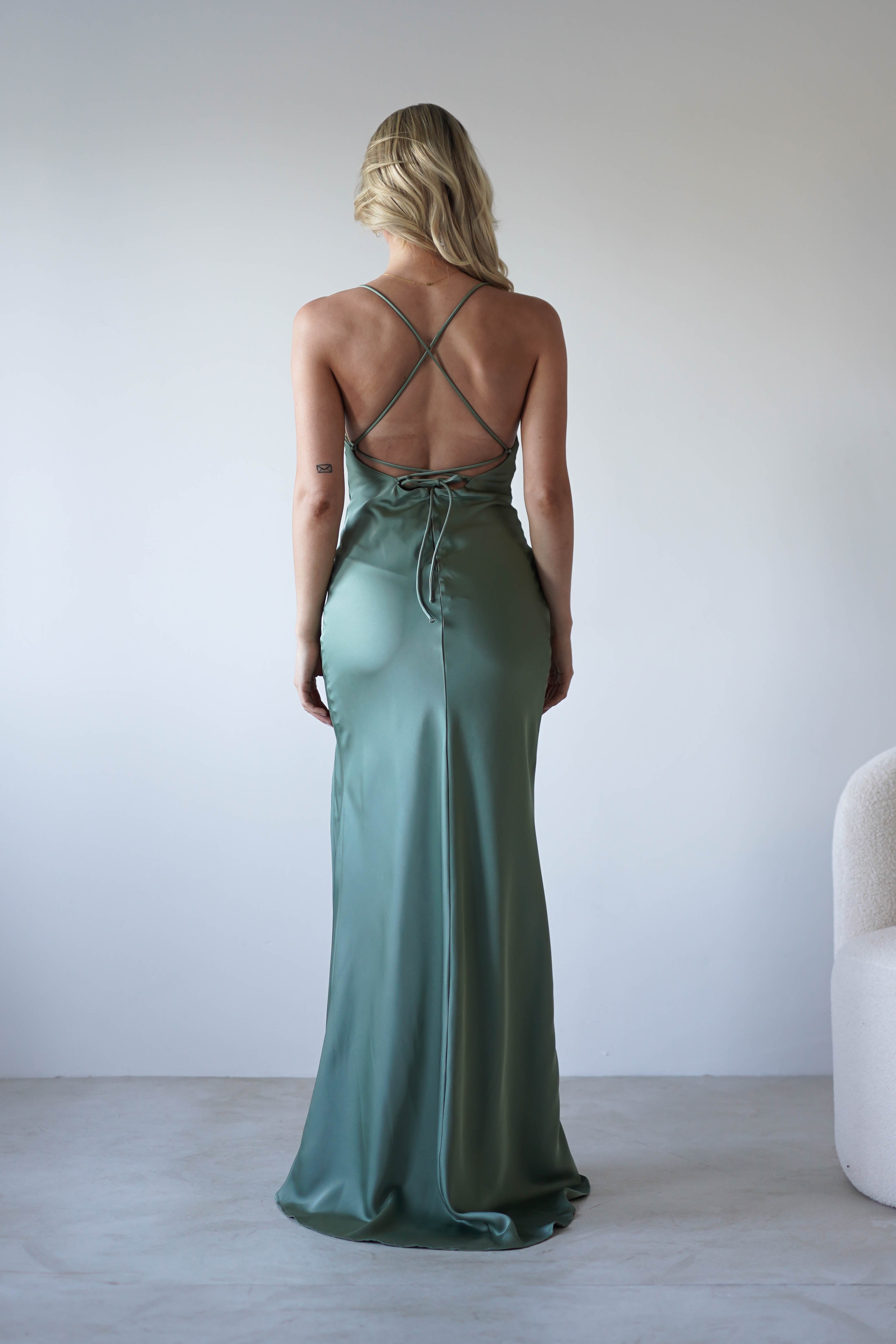 A woman with blonde hair, seen from behind, wears a long sage green satin slip dress with adjustable tie straps and an open back, standing in a minimalistic room with a light-colored chair.