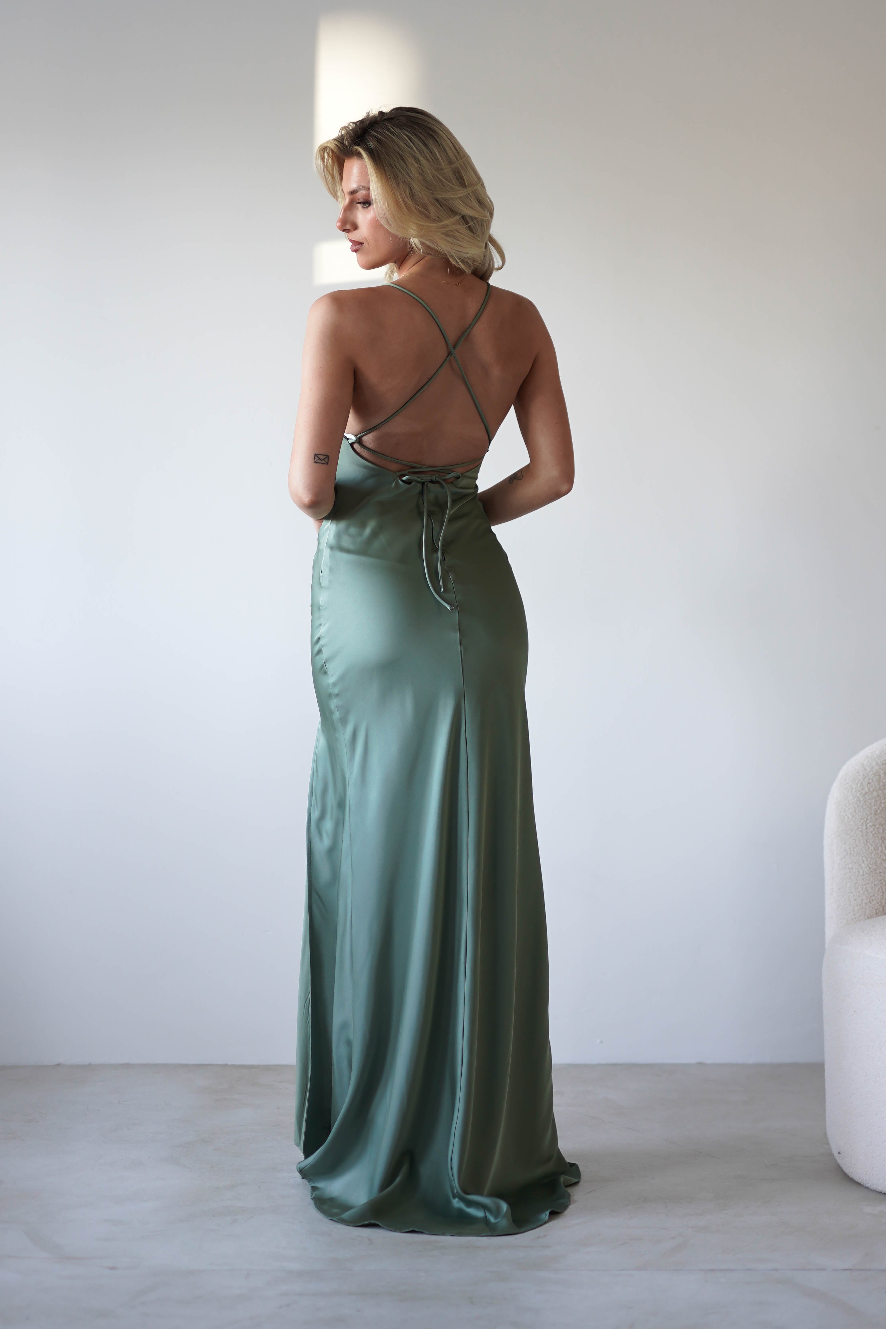 A woman in a sage green satin slip dress with adjustable tie straps and a crisscross back stands facing away in a minimalist room with light walls and a soft, curved chair.