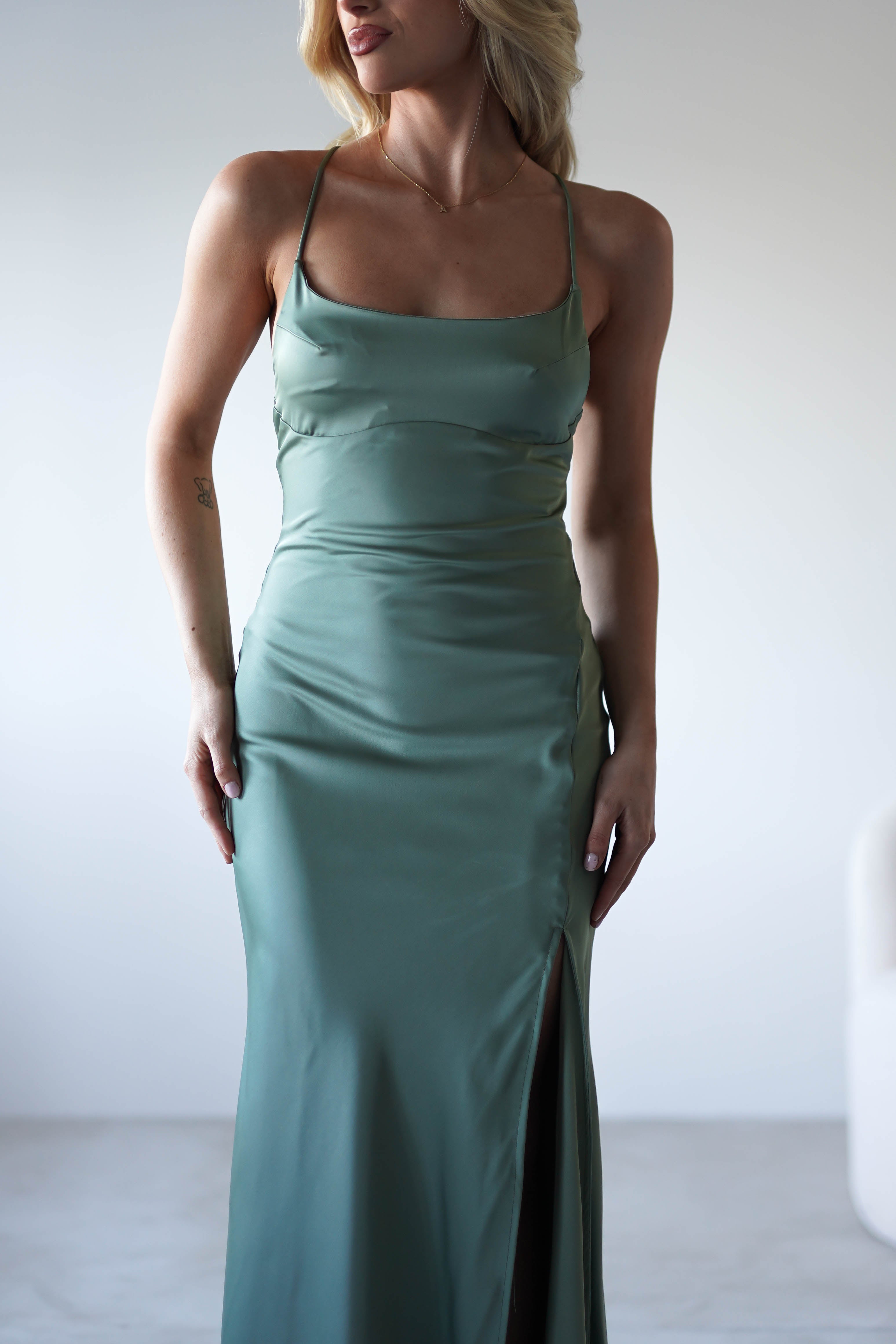 A woman wearing a fitted, sage green satin slip dress with adjustable tie straps and a thigh-high slit stands in a minimalist, light-filled room.