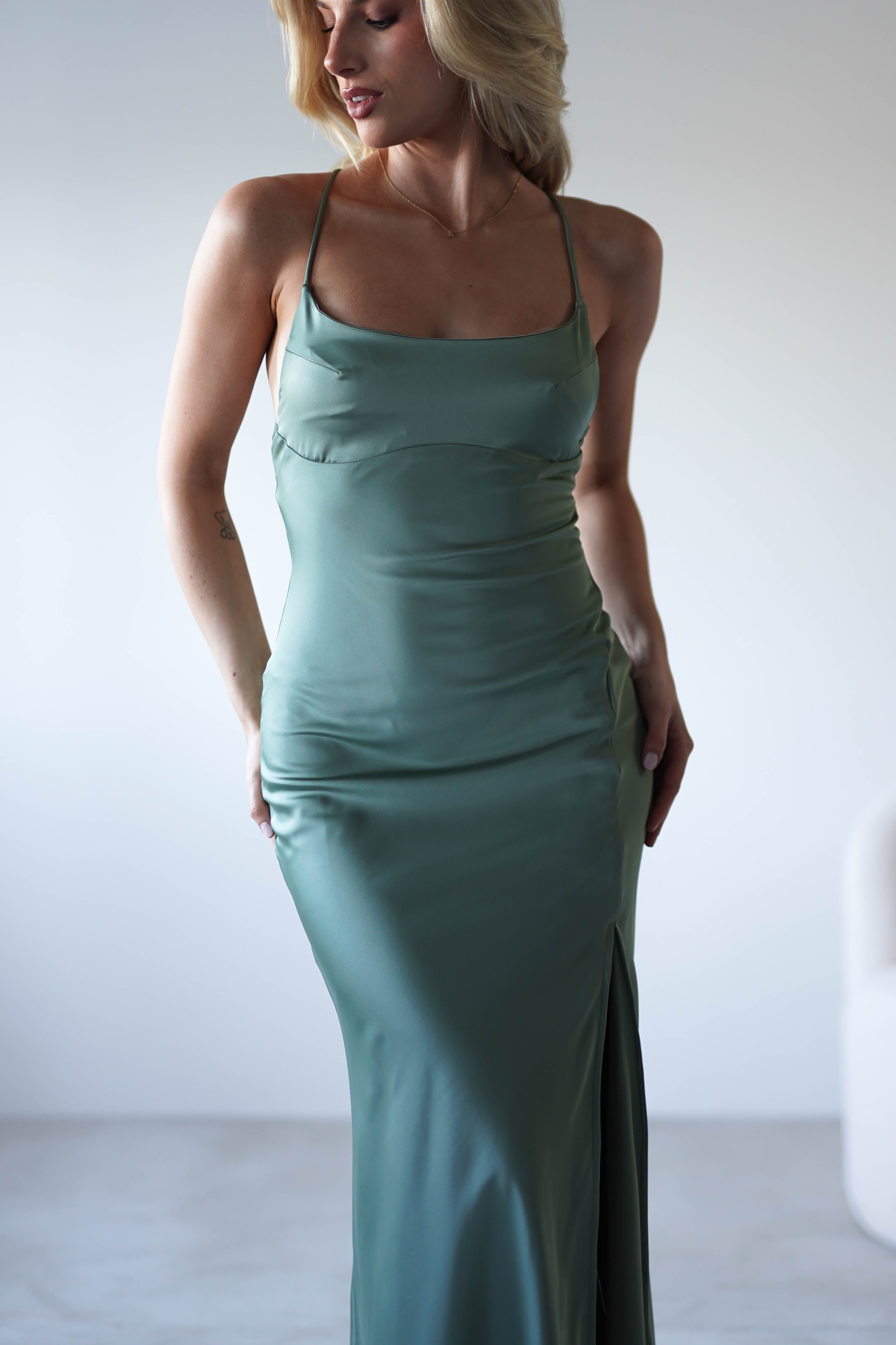 A woman stands indoors wearing a fitted, sage green satin slip dress with a thigh-high slit and adjustable tie straps. She poses with one hand on her hip against a minimal, softly lit background.