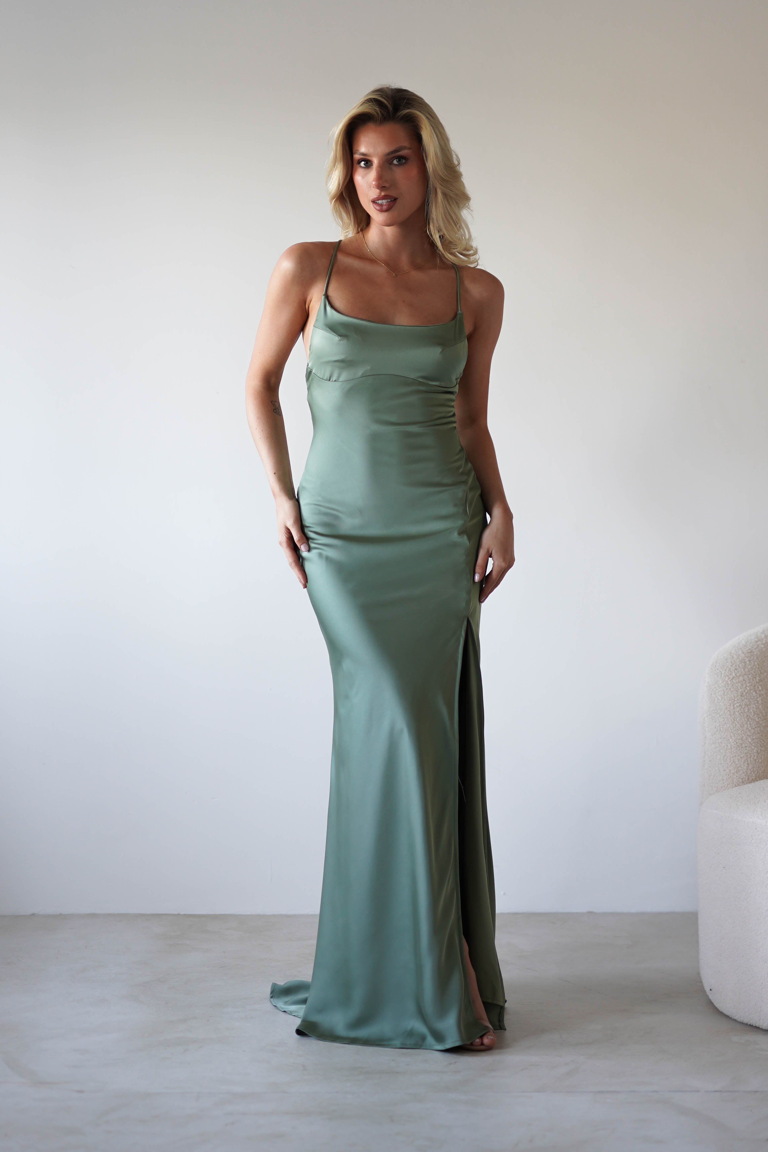 A woman stands indoors wearing a fitted sage green satin slip dress with adjustable tie straps and a thigh-high slit. She poses near a light-colored chair against a plain, light background.