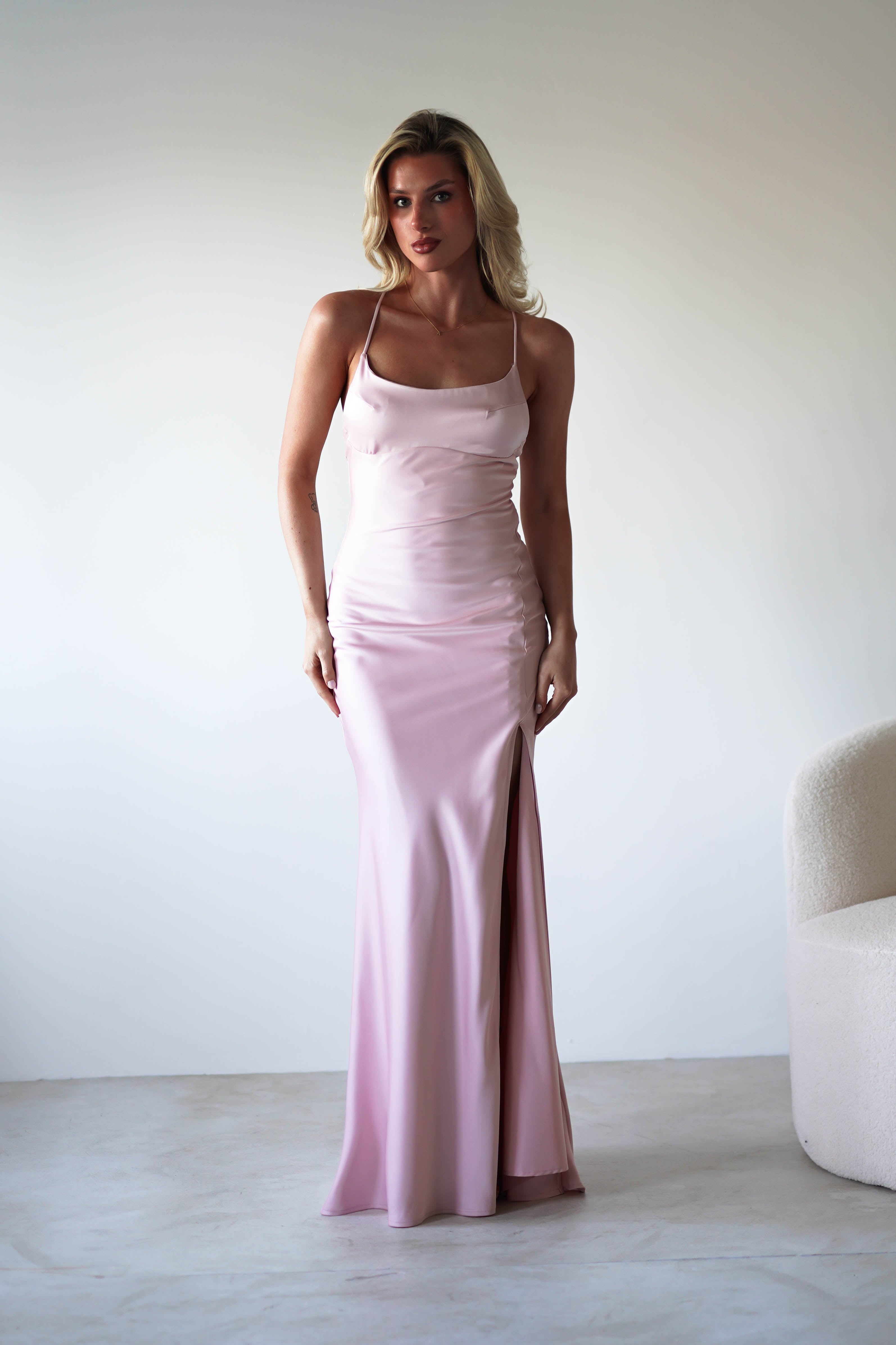 A woman with blonde hair wears a figure slimming slip dress in light pink sleek satin fabric, featuring a thigh-high slit and thin adjustable tie straps, standing indoors beside a curved white chair against a simple white background.