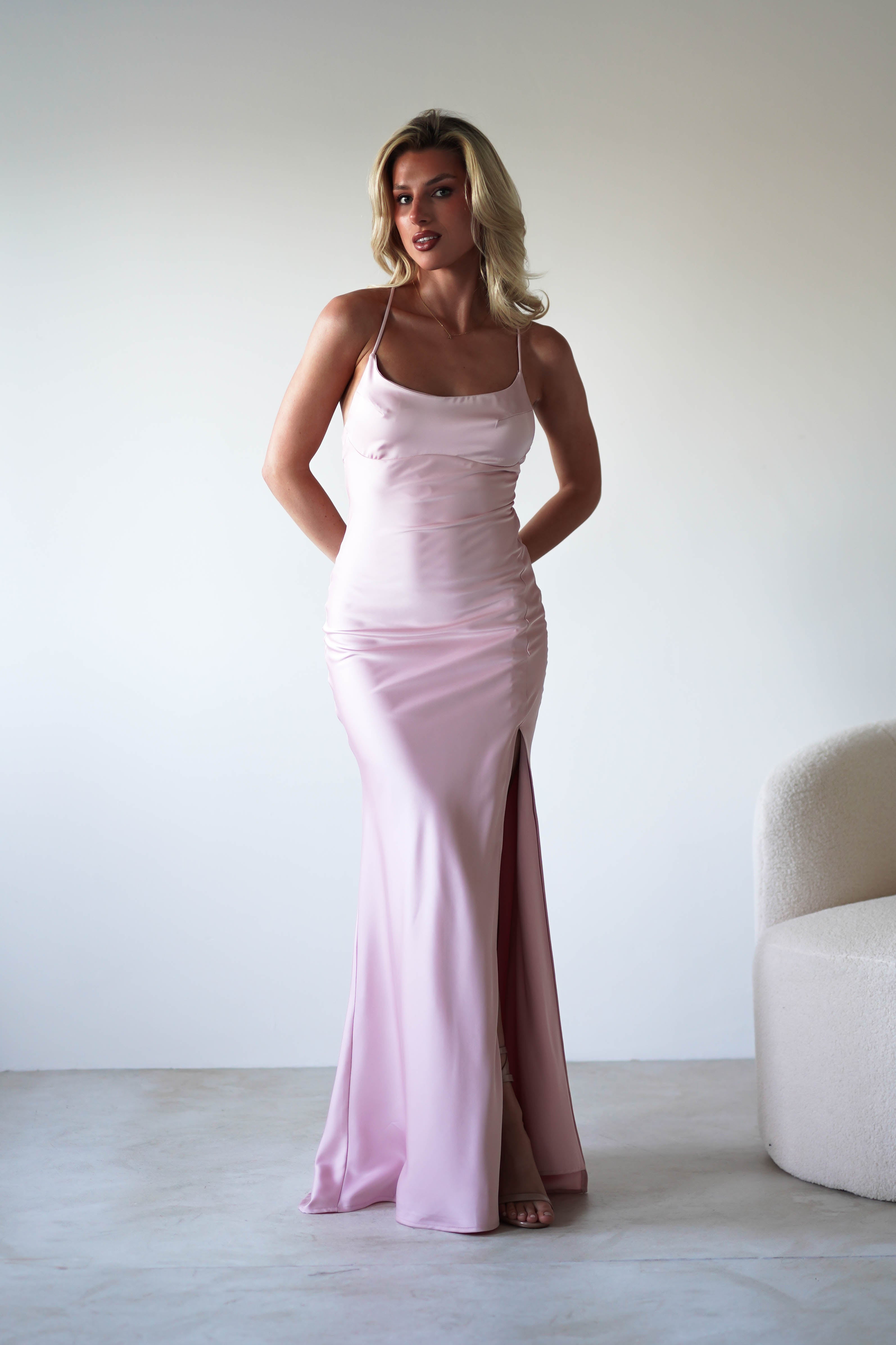 A woman in a light pink, floor-length figure slimming slip dress with sleek satin fabric and spaghetti straps stands indoors against a plain white wall, hands behind her back. The dress features a thigh-high slit. A cream-colored chair is partly visible on the right.