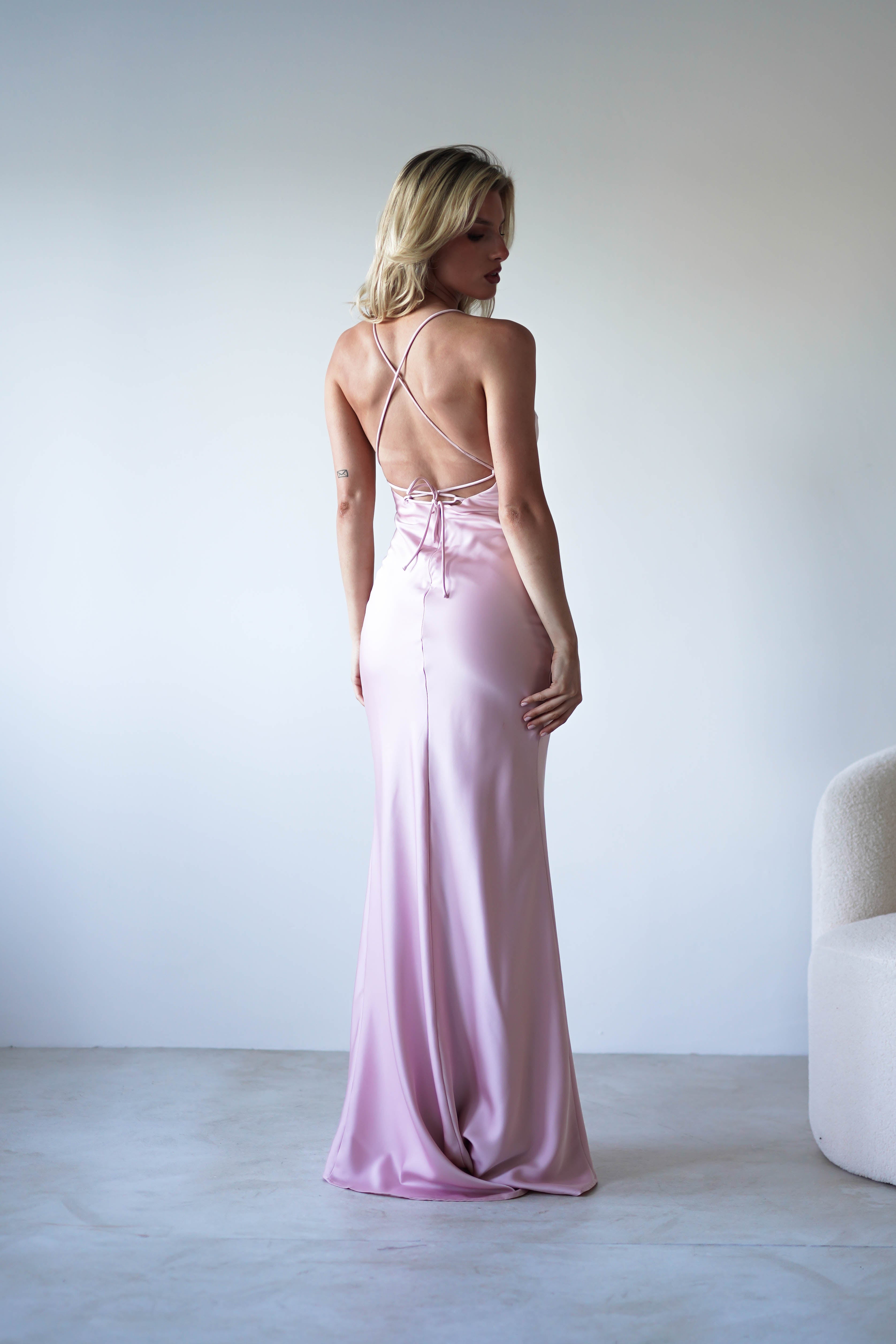 A woman stands facing away, wearing a long, figure slimming slip dress made from sleek satin fabric with crisscross adjustable tie straps. She is in a minimalist room with a white chair to the right and a light gray floor.