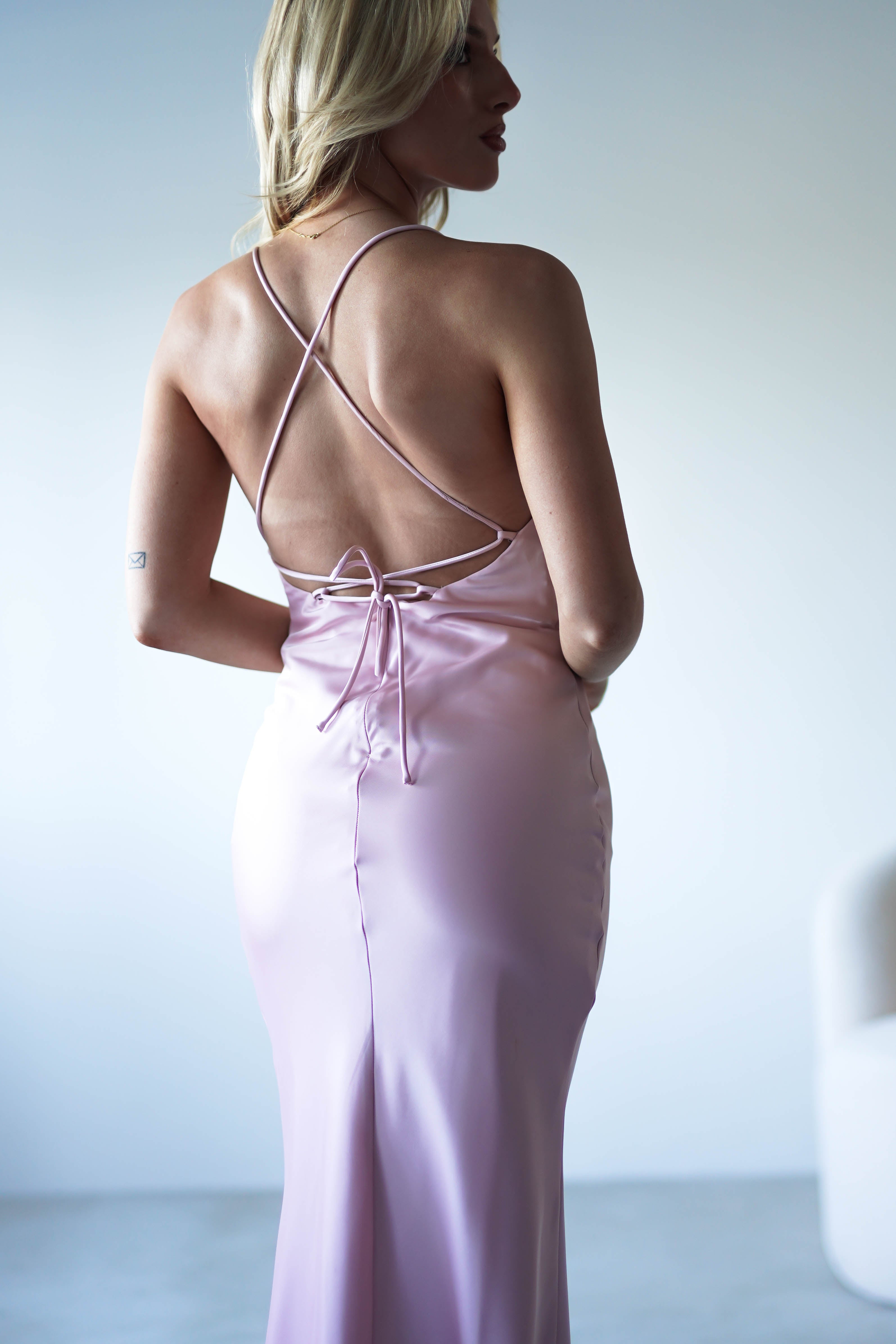 A woman with blonde hair is wearing a figure slimming slip dress in shiny, light pink satin with sleek satin fabric and thin crisscross straps, standing against a plain light background.