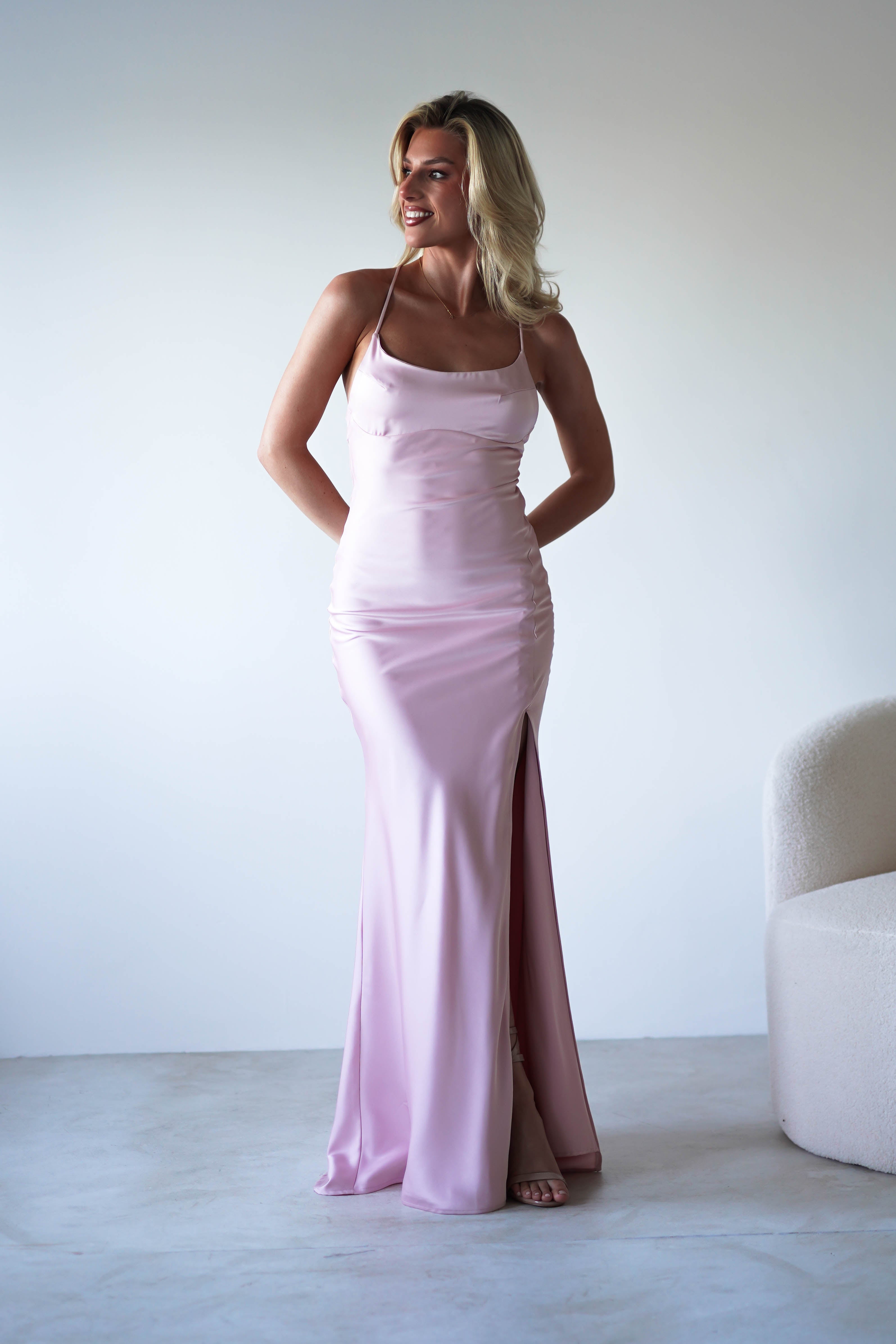 A woman in a pale pink, figure slimming slip dress with adjustable tie straps and a thigh-high slit stands smiling in a minimalist room next to a white textured chair.