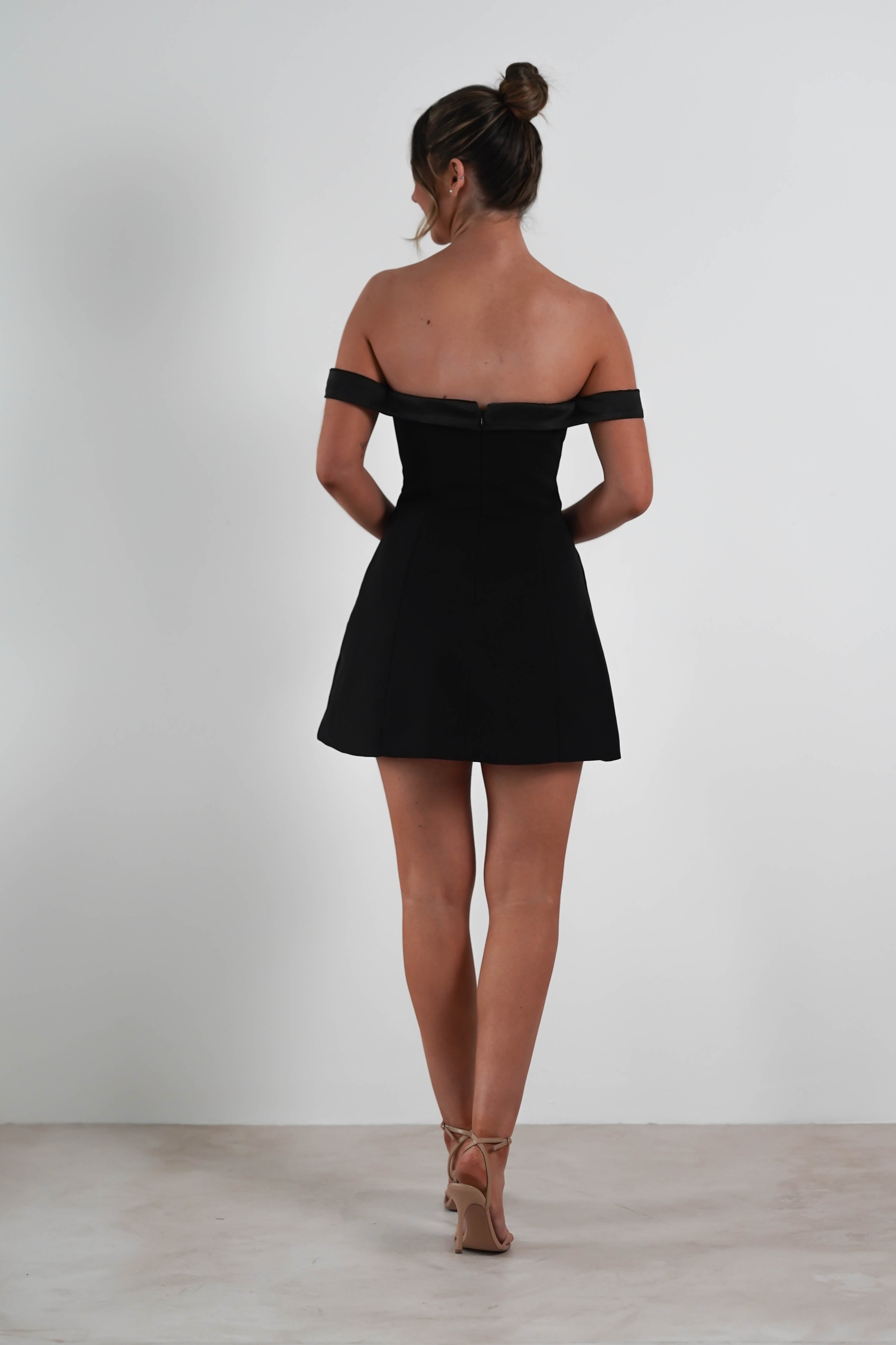 A woman, poised as a wedding guest, stands with her back turned in an off-the-shoulder black dress and nude heels. Her hair is styled elegantly in a bun against the plain white backdrop.