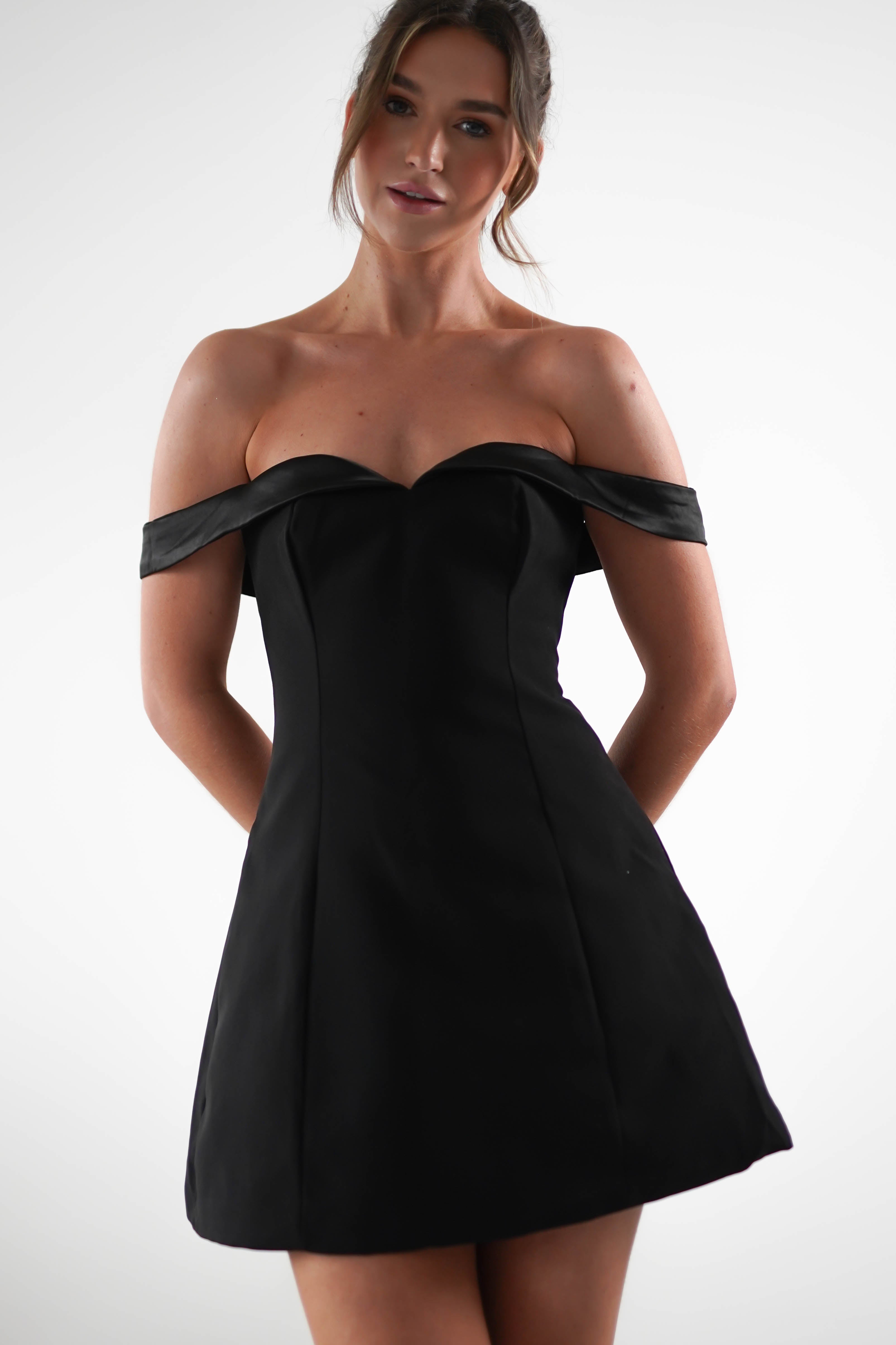 A woman in a strapless black dress, perfect for a wedding guest, stands against a plain white background with a soft expression. Her hair is styled in a loose updo, complementing the elegant off-the-shoulder look.
