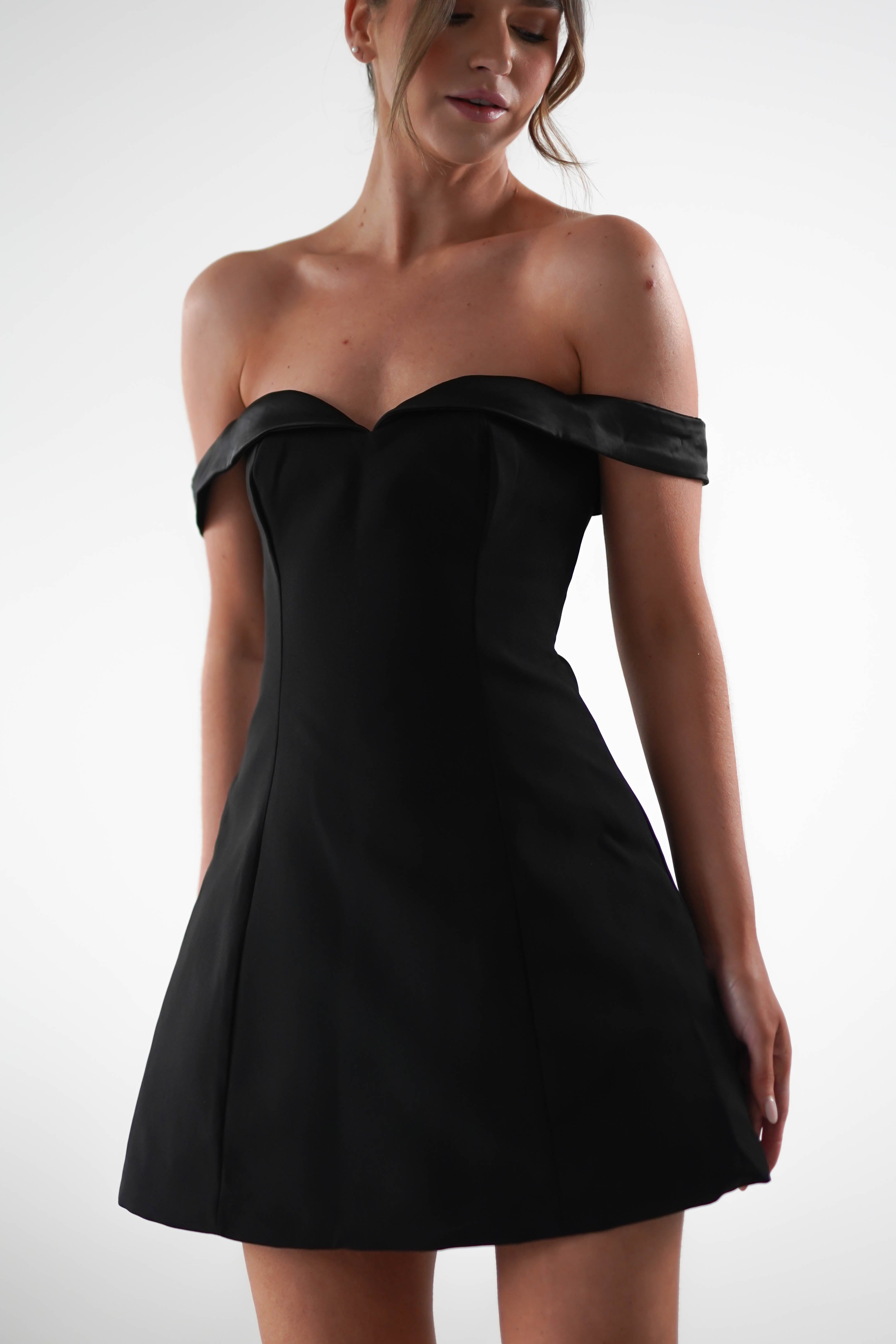 A woman showcases an elegant off-the-shoulder black dress, perfect for a wedding guest. Its design features a fitted bodice and a flared skirt, exuding timeless charm as she stands against a plain, light background.