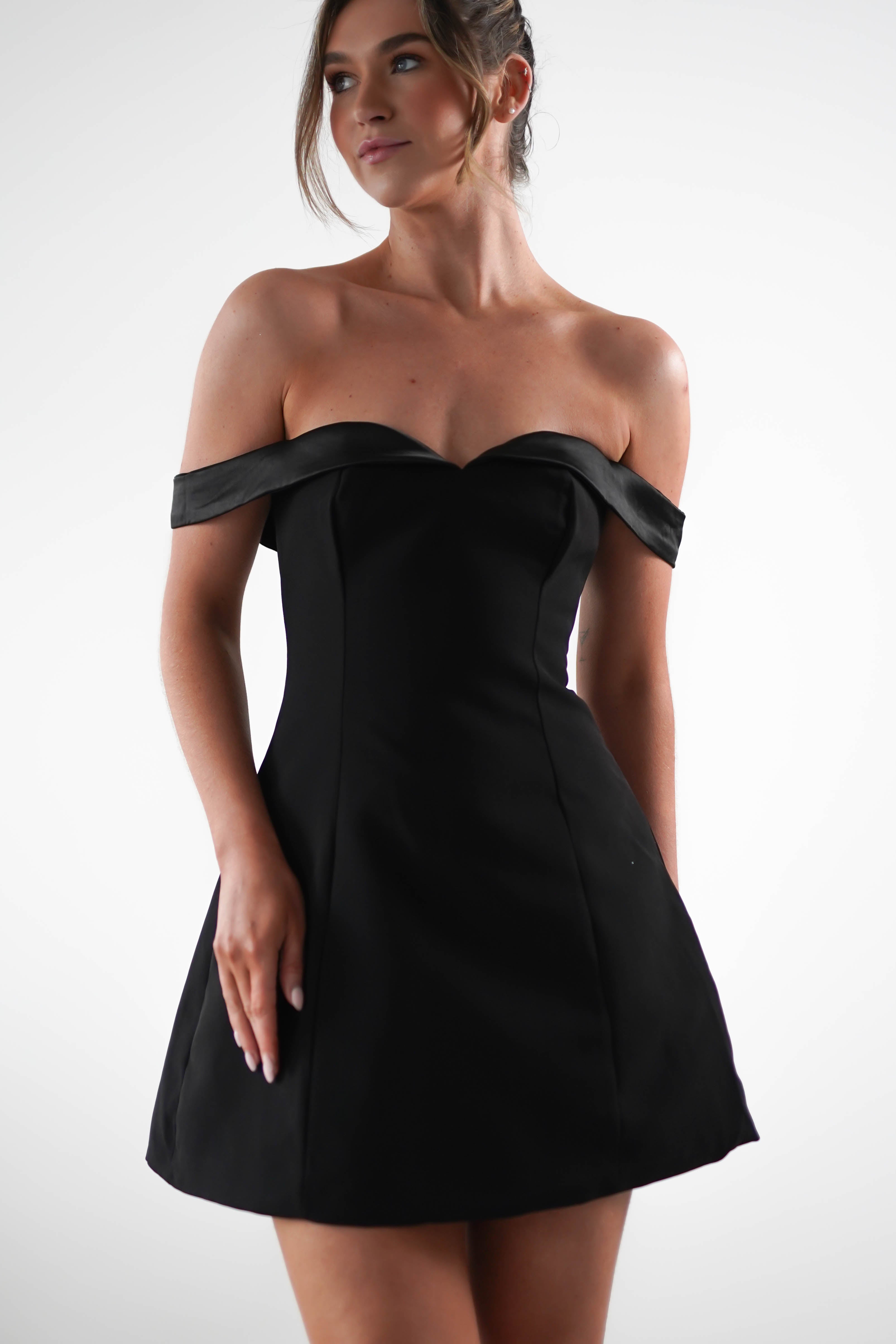 A person wearing a black off-the-shoulder dress, perfect for a stylish wedding guest, stands against a plain white background. The knee-length dress with a fitted bodice complements their up-styled hair as they look slightly to the side.