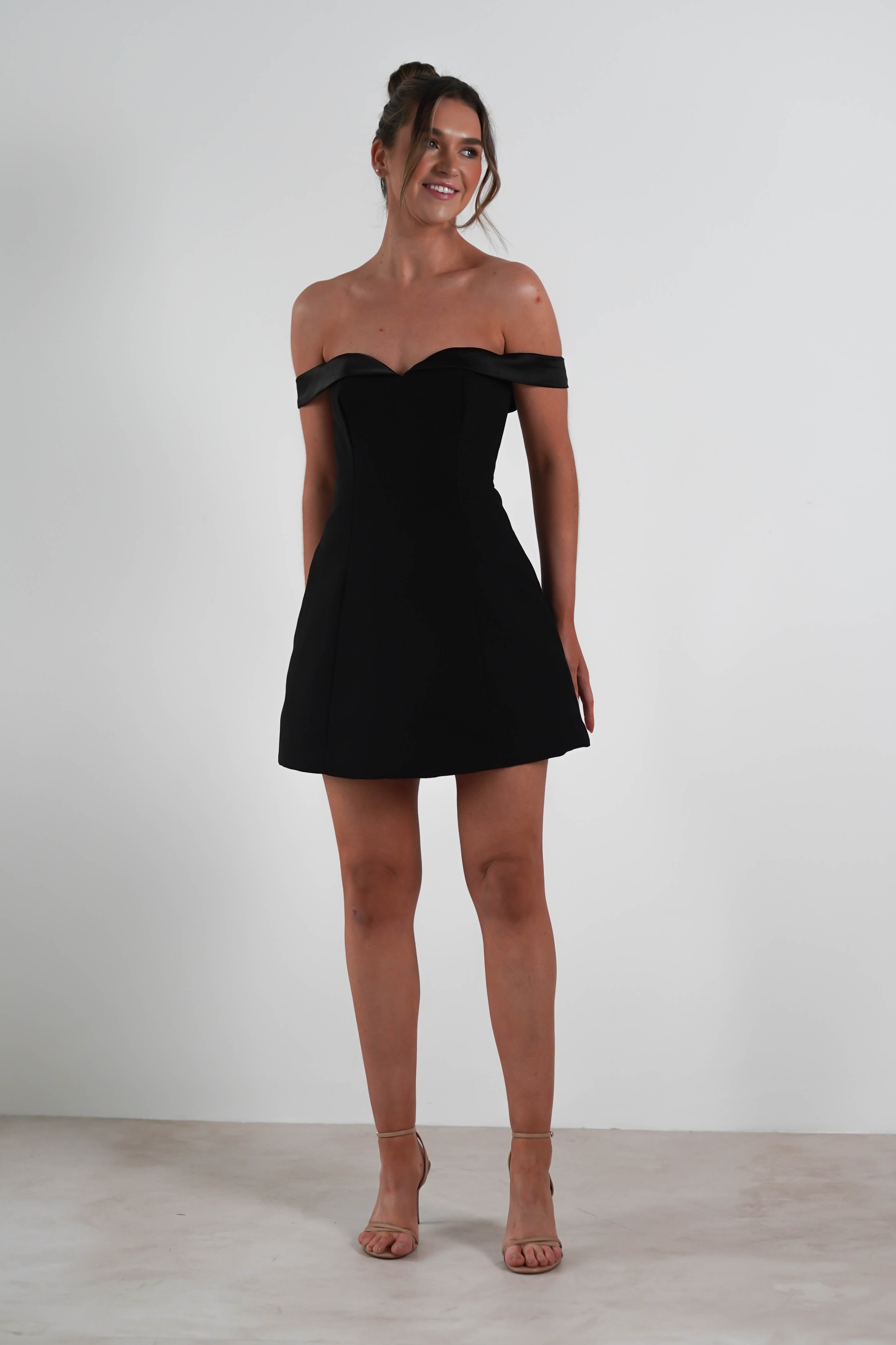 A person in an off-the-shoulder black dress, perfect for a wedding guest, stands against a plain white background. Their hair is tied up and they are smiling, with one leg slightly bent. They complete the look with beige high-heeled sandals.