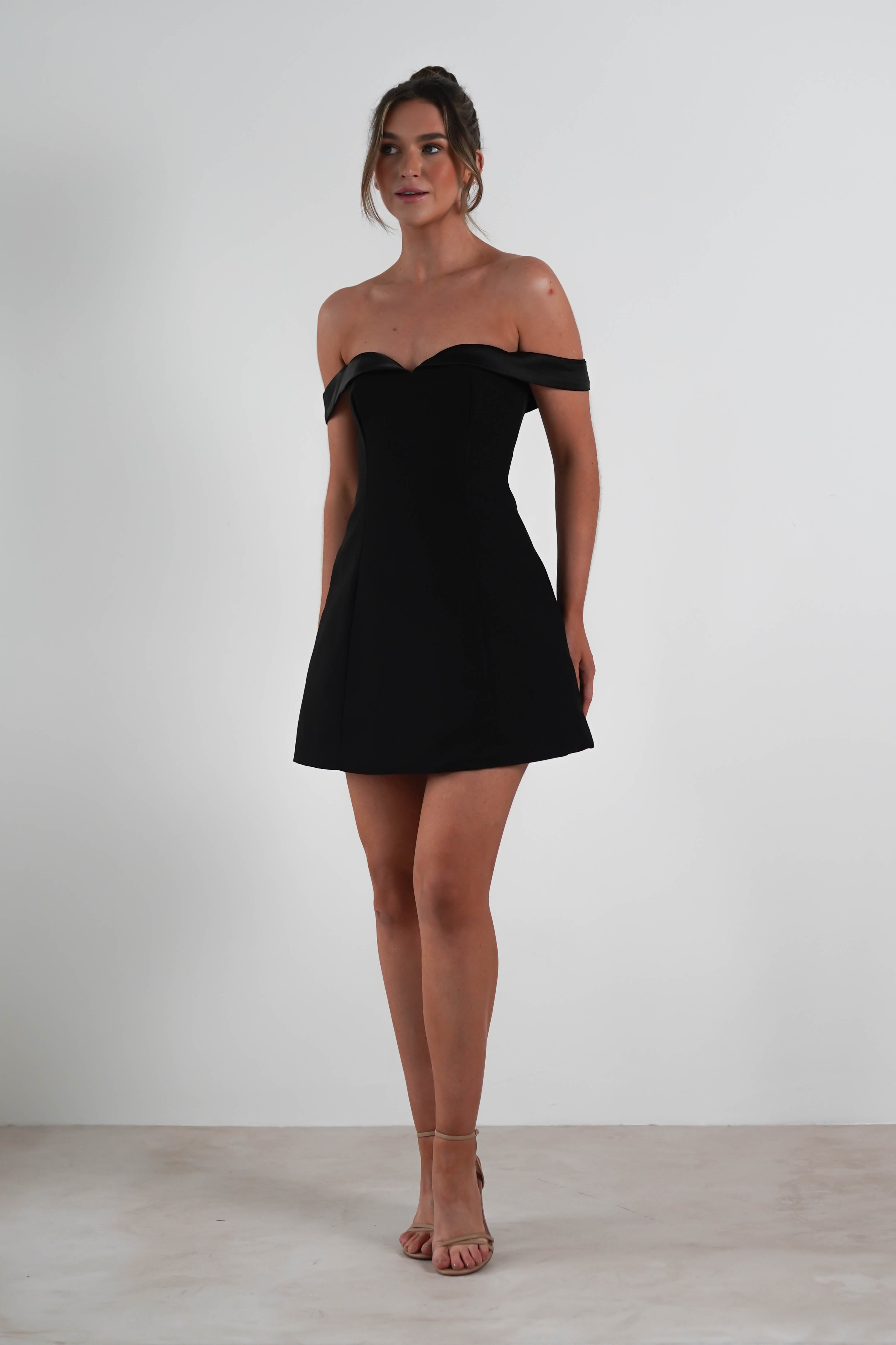 A woman wearing a stylish off-the-shoulder black dress stands against a plain white background. The sweetheart mini design complements her neutral expression, and her hair is styled up. She completes the look with nude heels, standing gracefully with her arms relaxed at her sides.